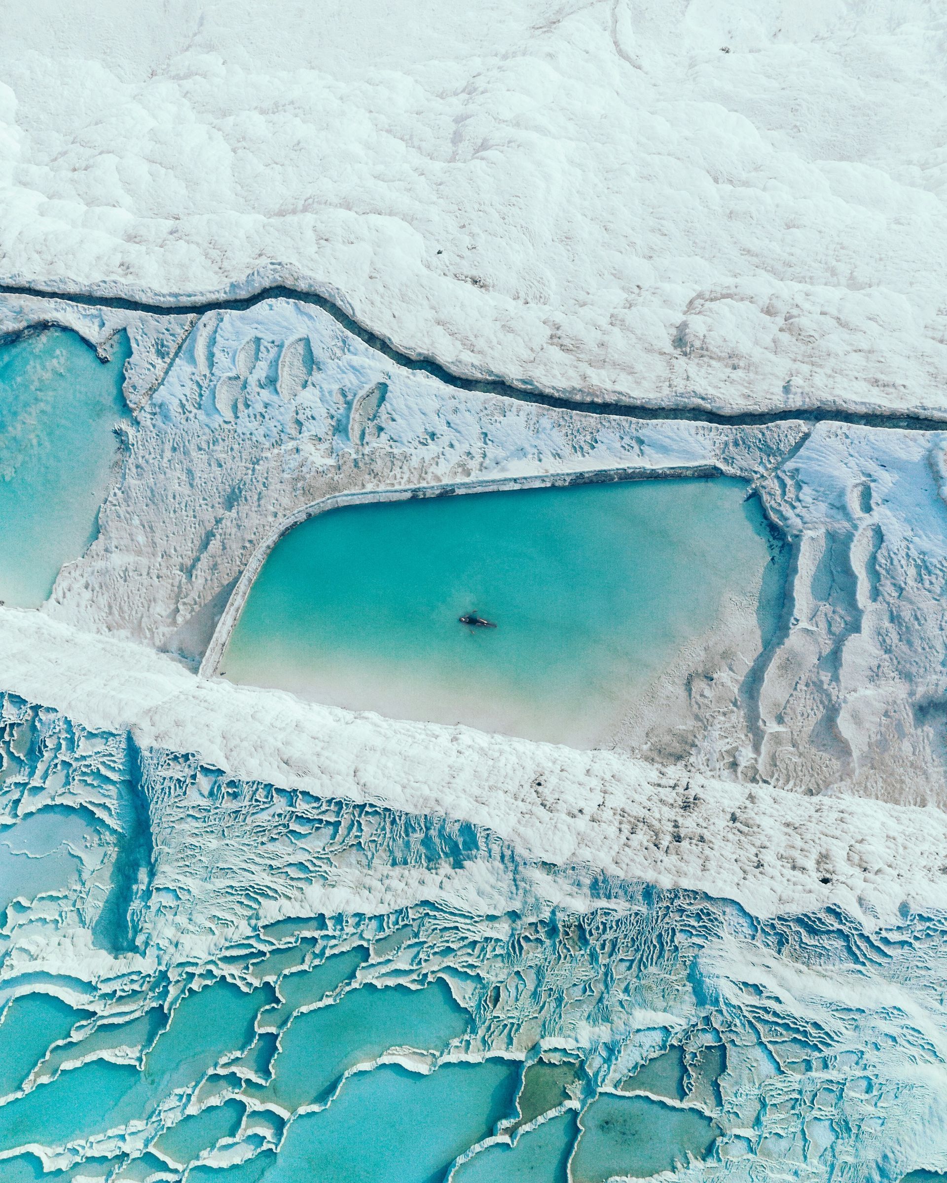 Aerial view of turquoise thermal pools in white travertine terraces, with one person swimming in Pamukkale, Turkey.