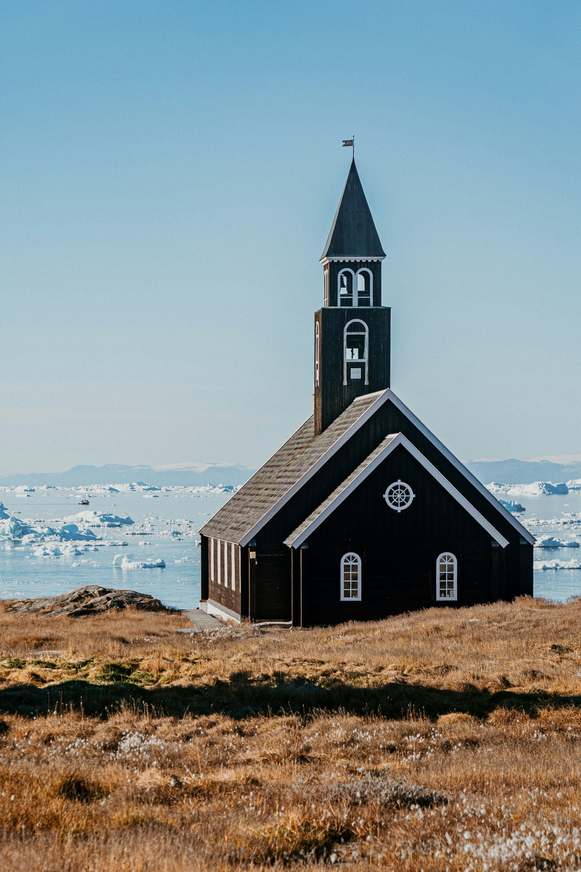 Black church with a steeple overlooking a frozen sea, under a clear blue sky at the Ilulissat Icefjord, Greenland.