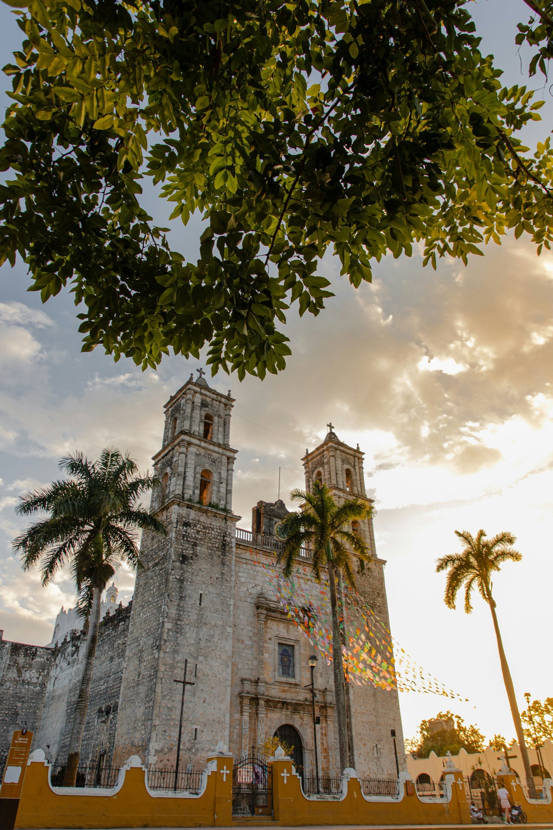 Old stone church with two bell towers with palm trees and a cloudy sky at sunset in front in Valladolid, Mexico.