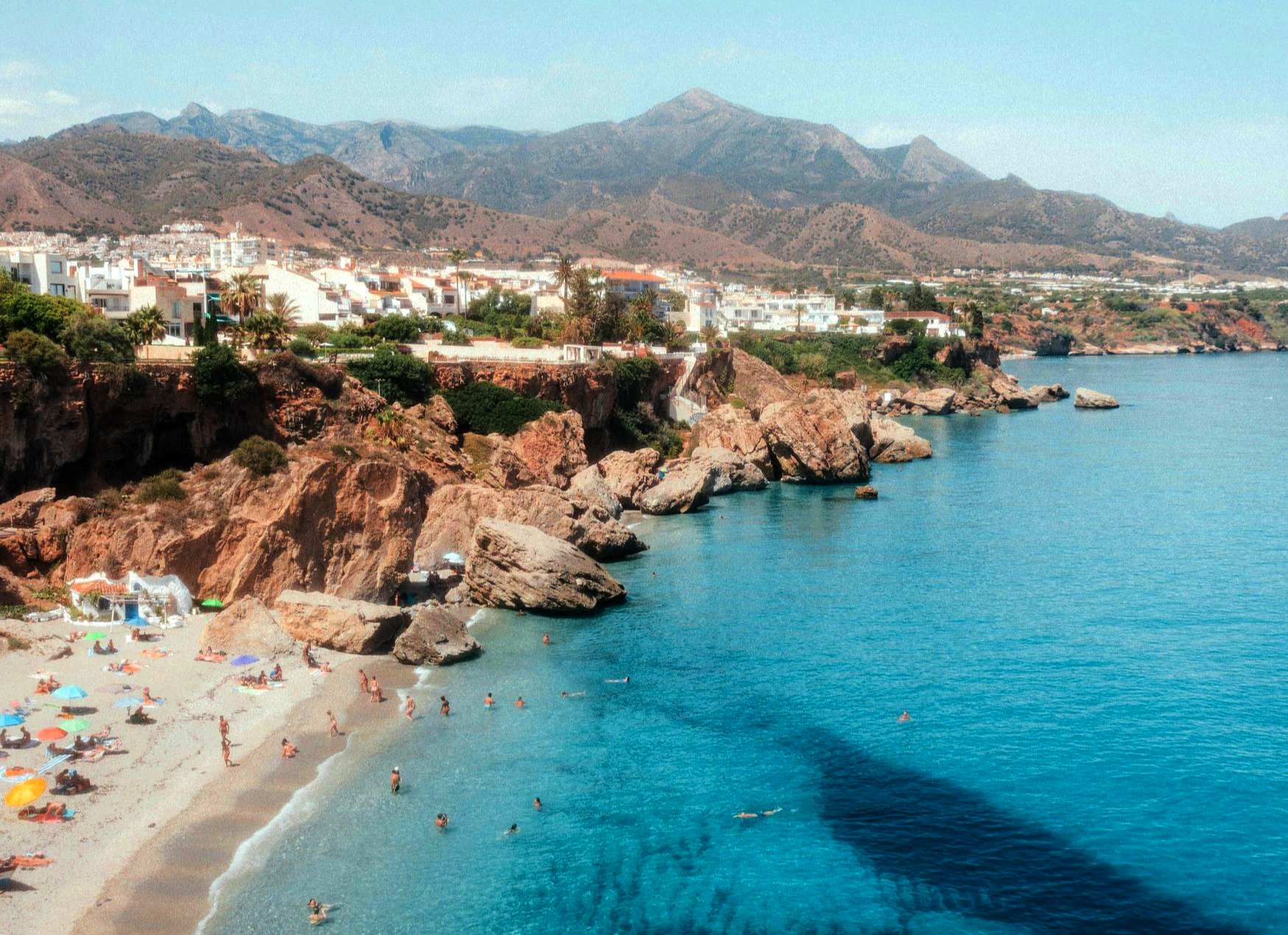 Beach with clear turquoise water, rocky cliffs, and a town with white buildings at the mountains' base in Spain.