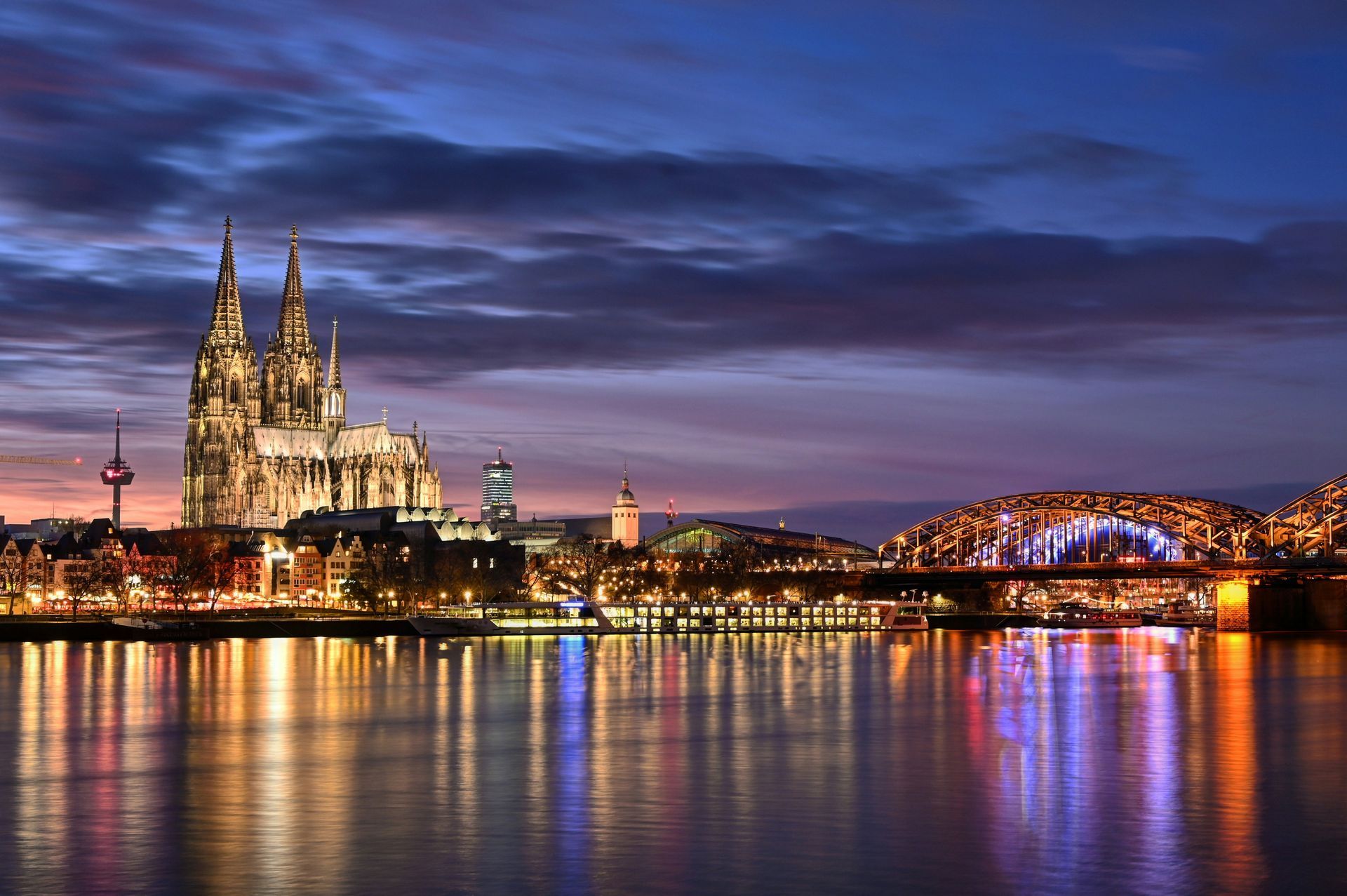 Cologne skyline at dusk, with illuminated cathedral, bridge, and river reflecting city lights.