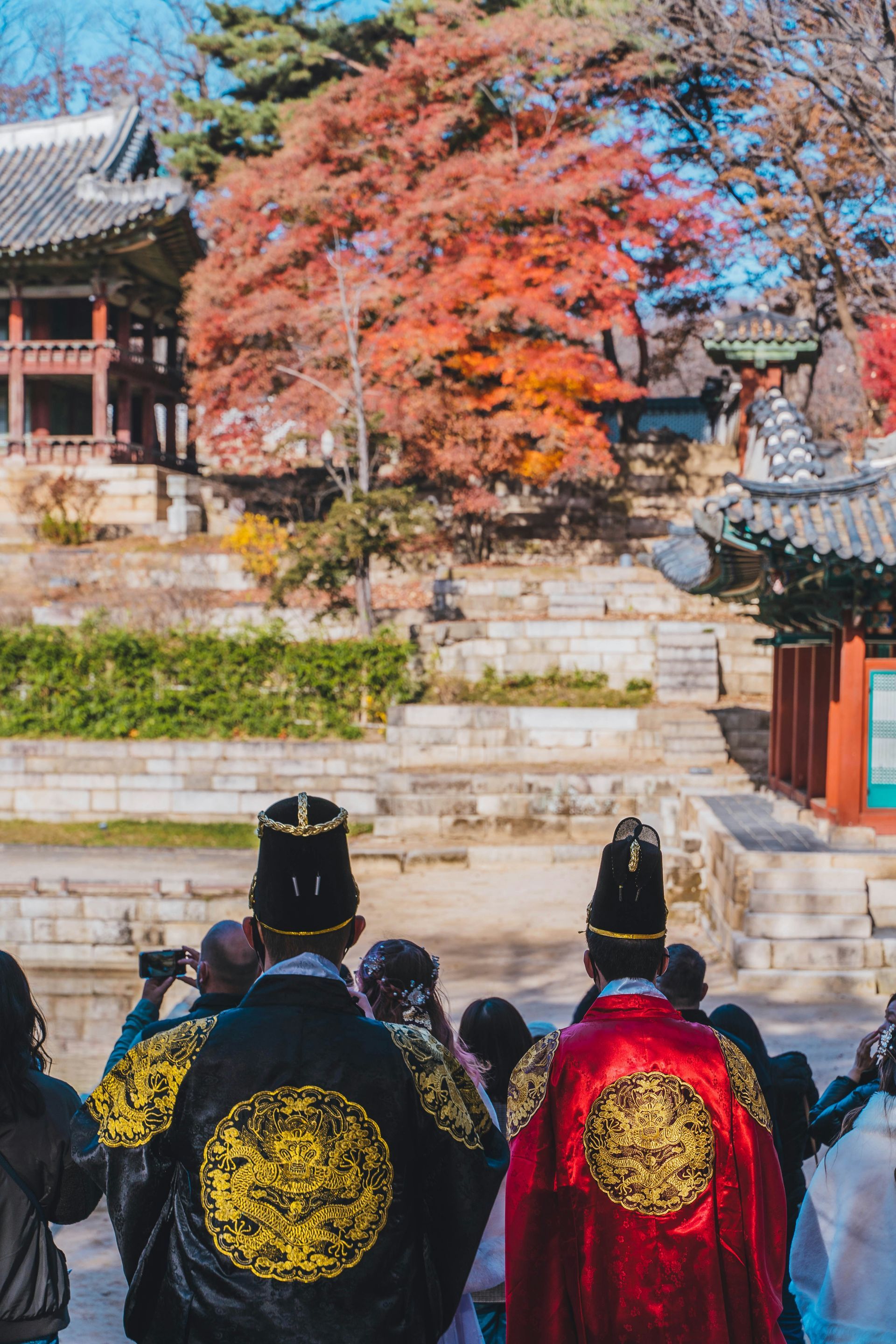 Two people in traditional Korean royal attire, facing away. Autumn foliage and a traditional building in the background.