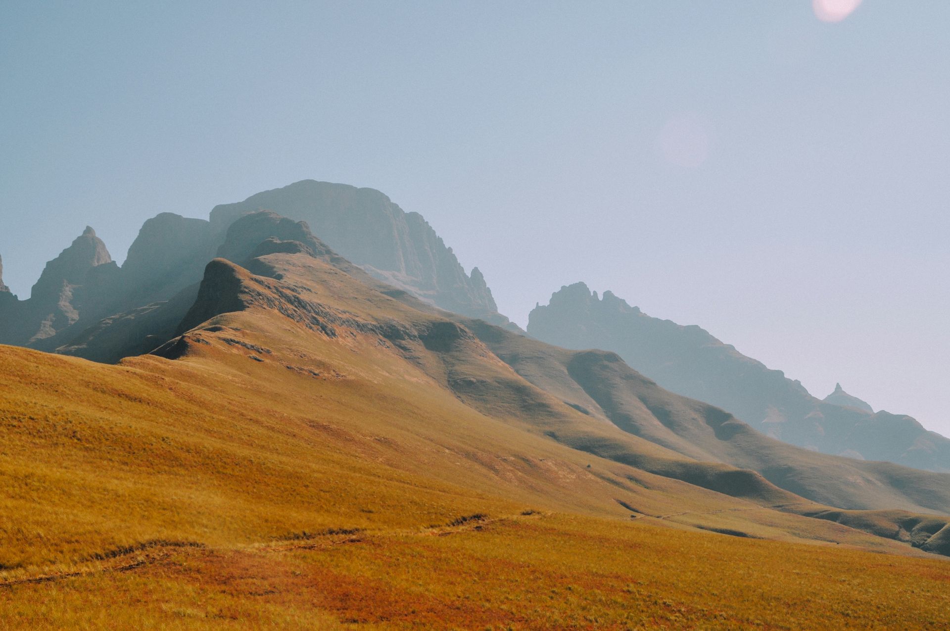 Golden field sloping up to a mountain range under a hazy blue sky in the Drakensberg, South Africa.
