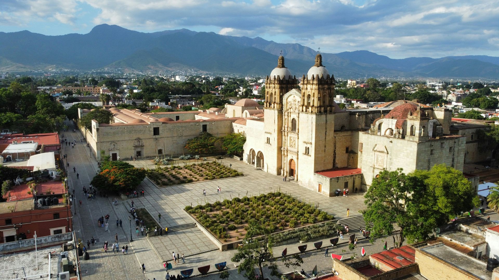 An aerial view of the golden-hued Santo Domingo church in a plaza with mountains in the background in Oaxaca, Mexico.