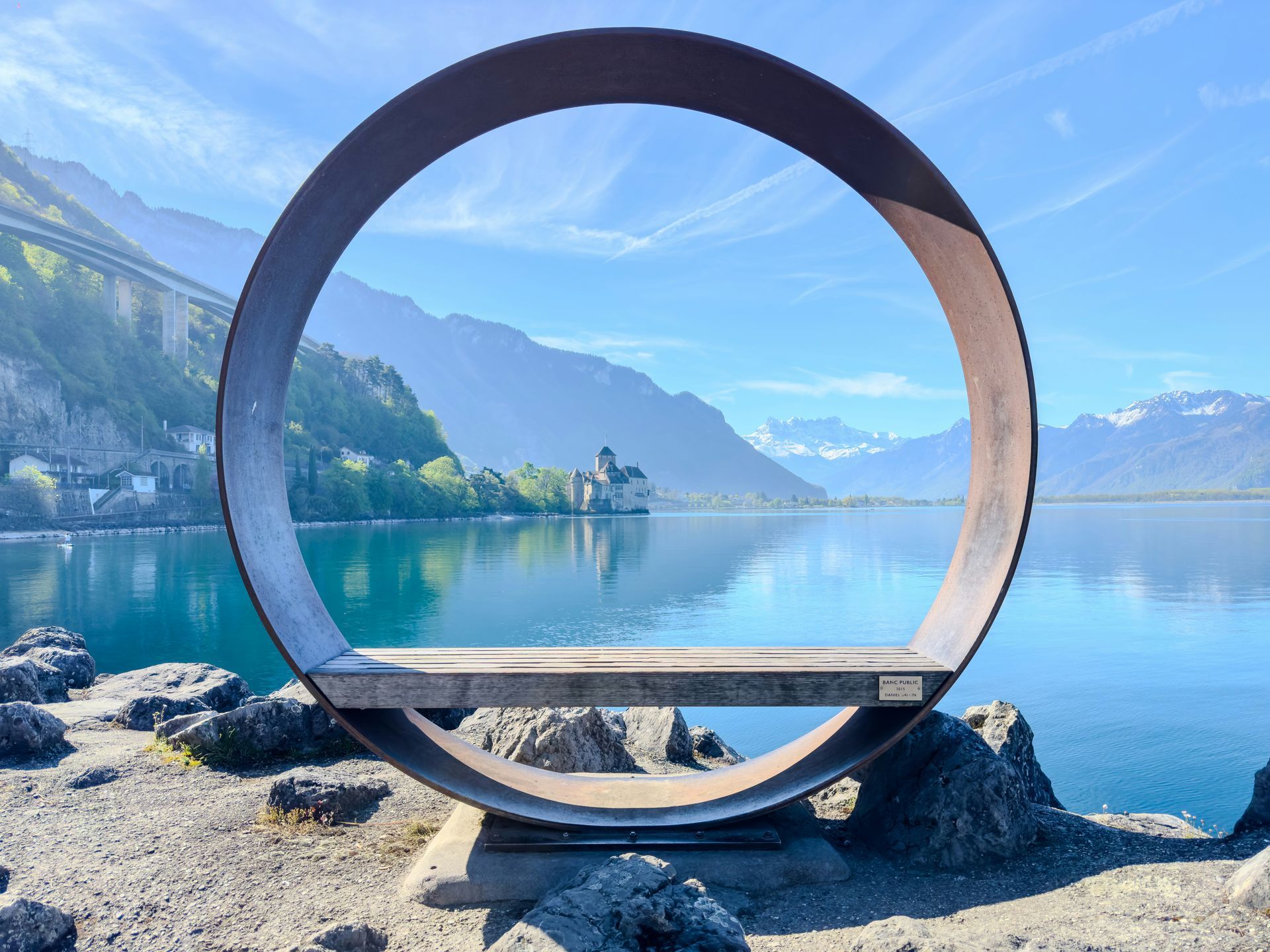 A large metal circle with a bench frames a lake, castle, and mountains in Montreux, Switzerland.