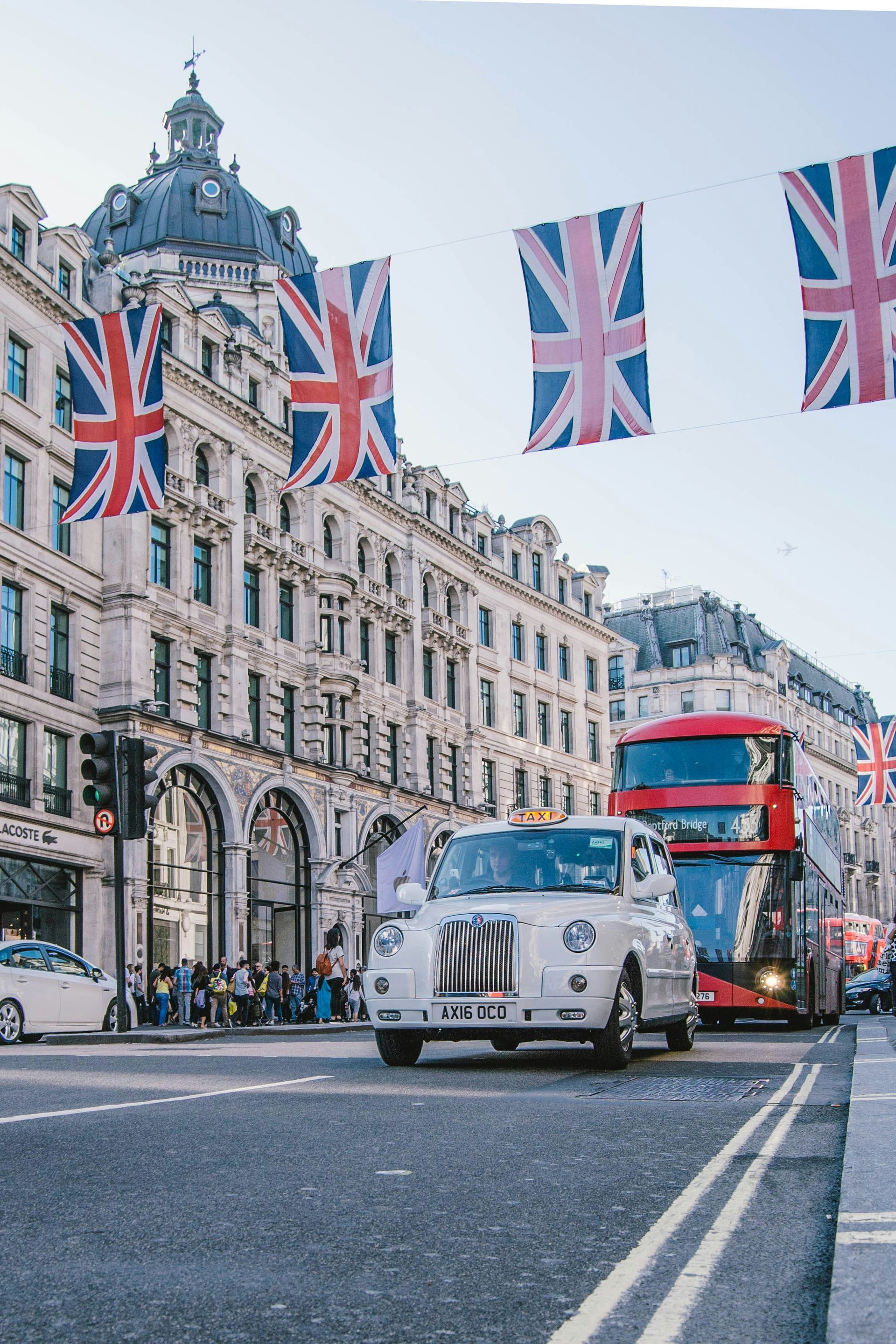 White taxi and red double-decker bus travel on a London street, Union Jack flags hang above.