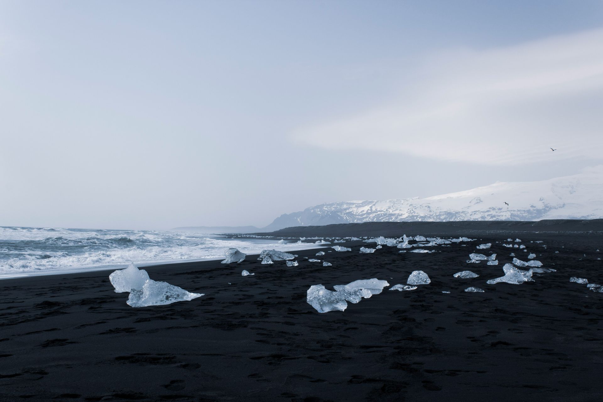 Black sand beach with scattered ice chunks, waves, and snow-covered mountains at Diamond Beach, Iceland.