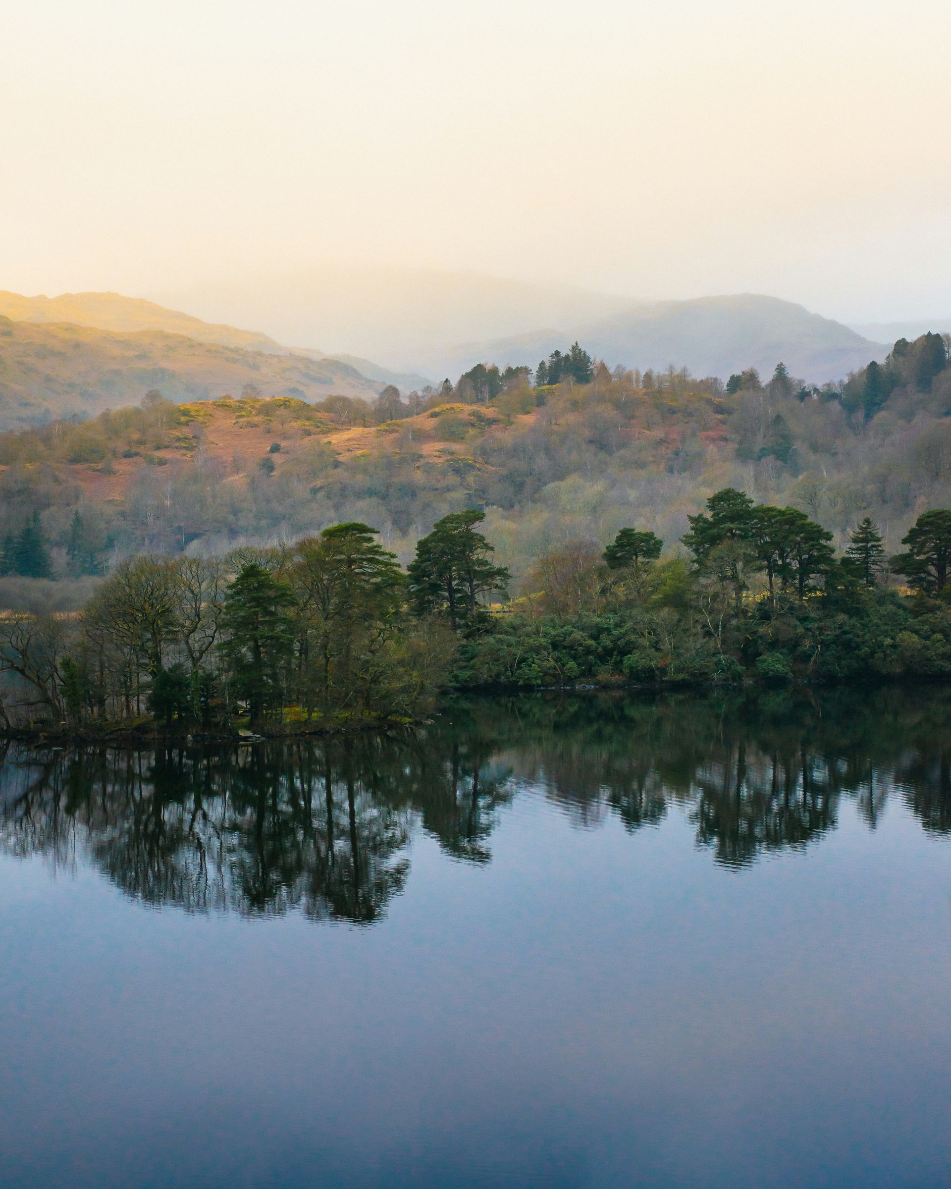 Trees reflected in calm water, with hazy hills and a pale sky in the background in the Lake District, England.