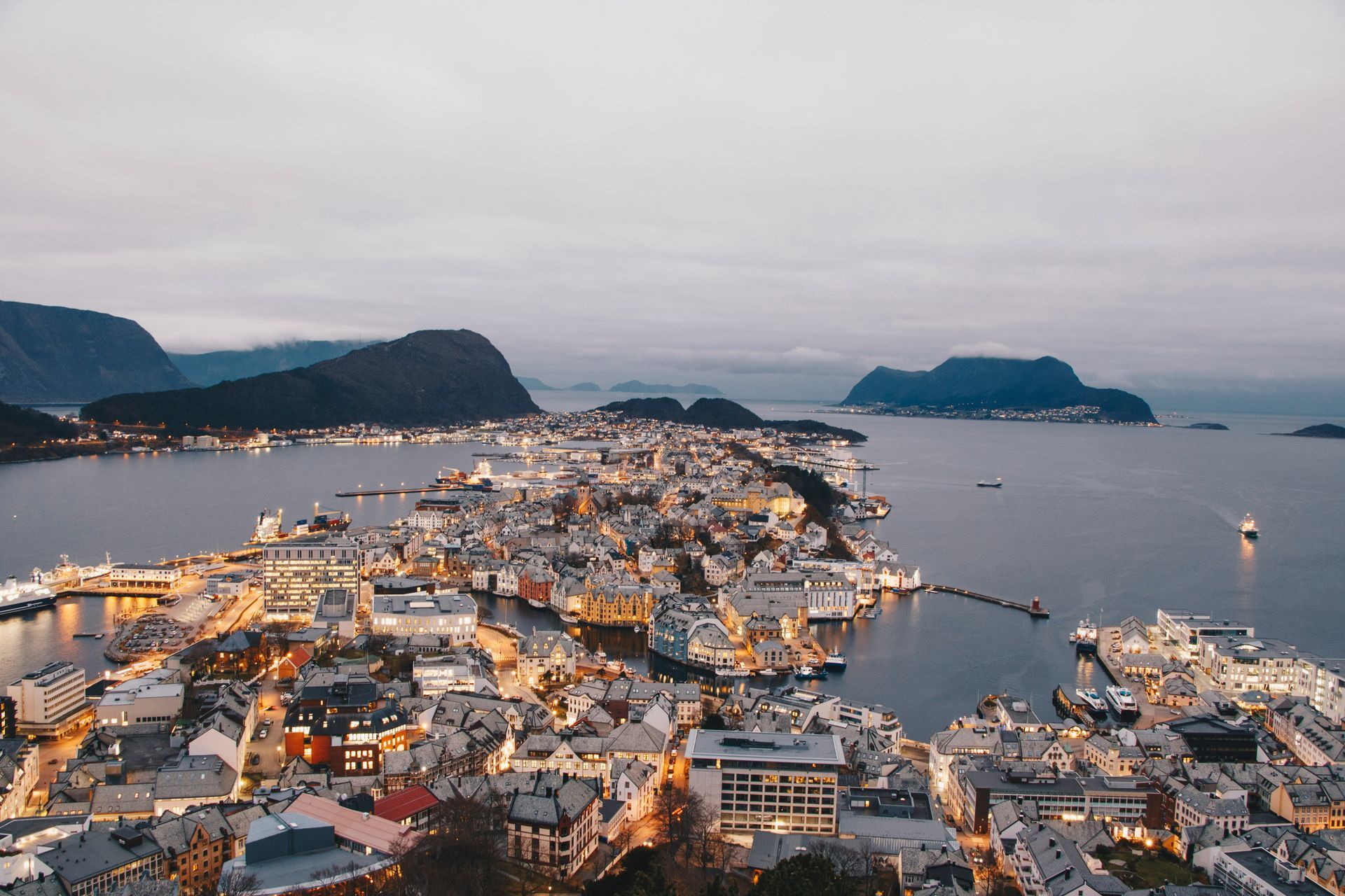 Cityscape at dusk with lights illuminating buildings and waterways, mountains in the background in Ålesund, Norway.