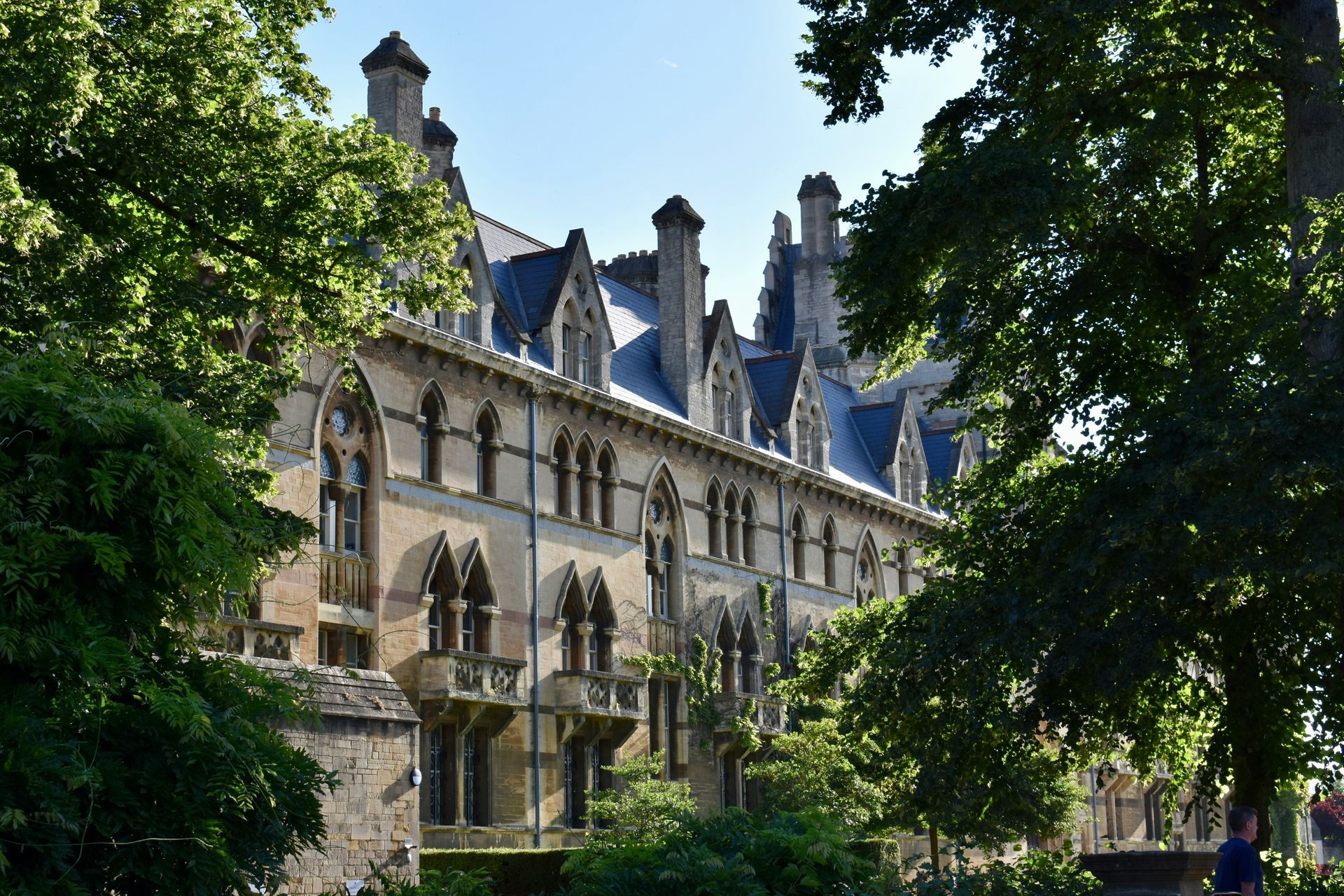Stone building with arched windows and chimneys, framed by green trees under a blue sky at Oxford University, England.