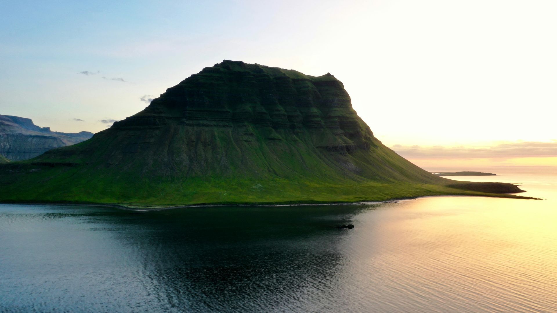 Green mountain rises from calm water at dawn in Snæfellsnes, Iceland.
