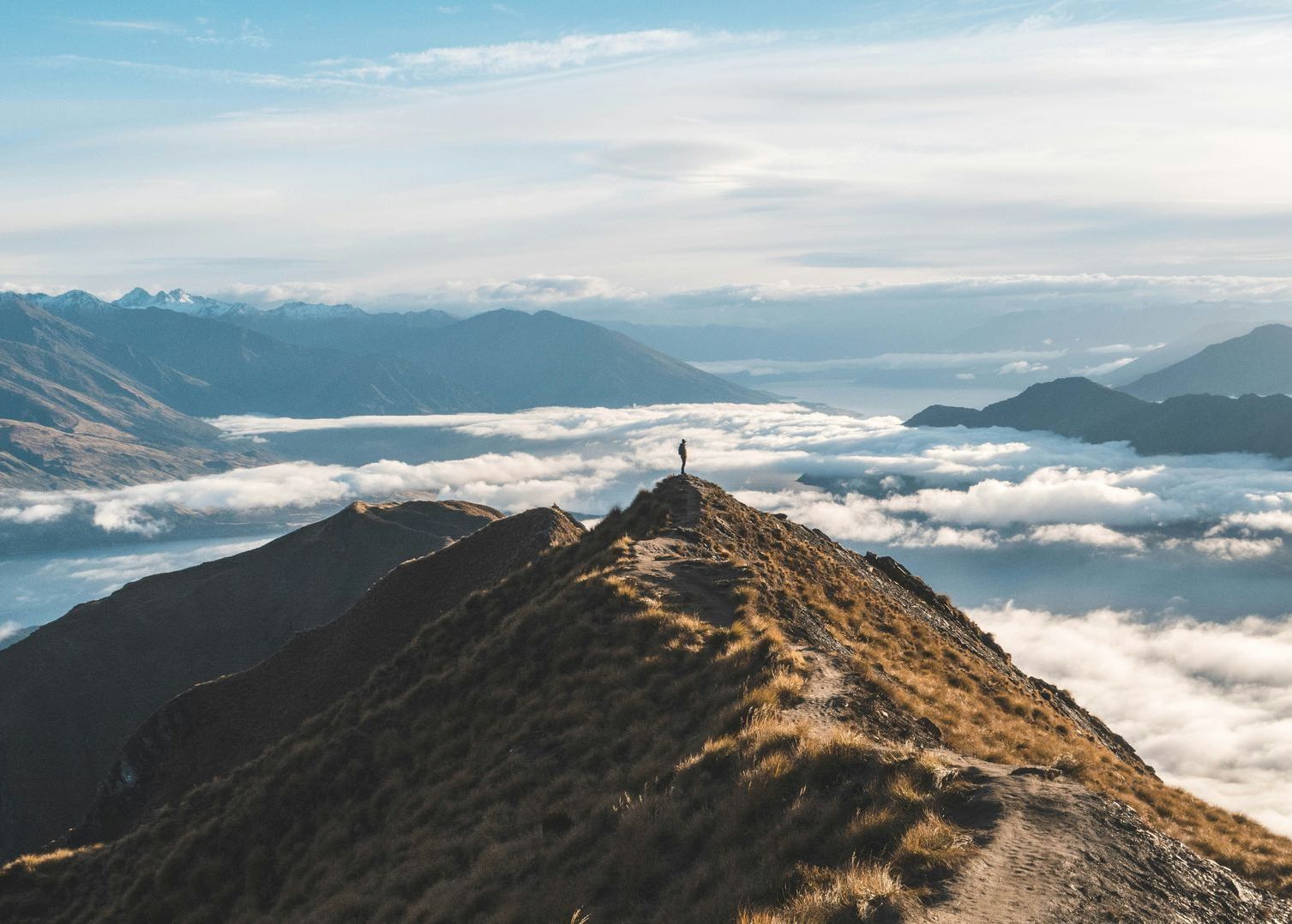 A person is standing on top of a mountain surrounded by clouds in New Zealand.