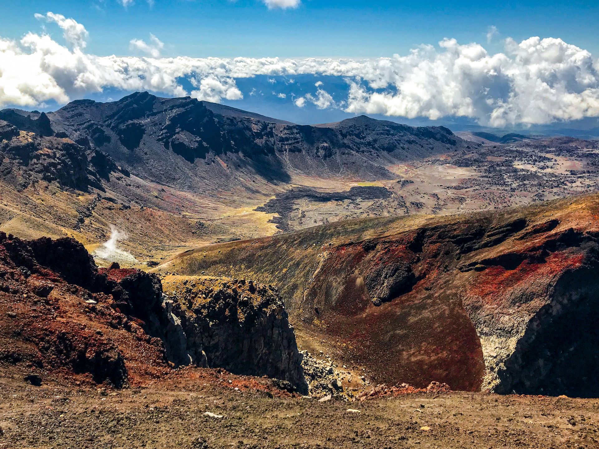 A view of a valley surrounded by mountains and clouds from the top of a mountain in Tongariro National Park, New Zealand.