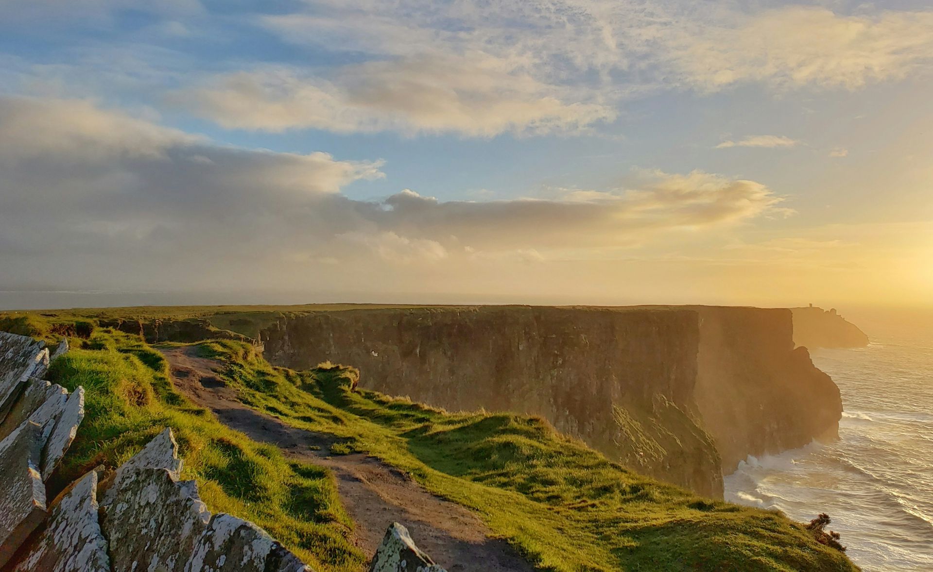 Cliffs of Moher at sunset. Ocean waves crash against the cliffs, with golden sunlight and green grass in Ireland.