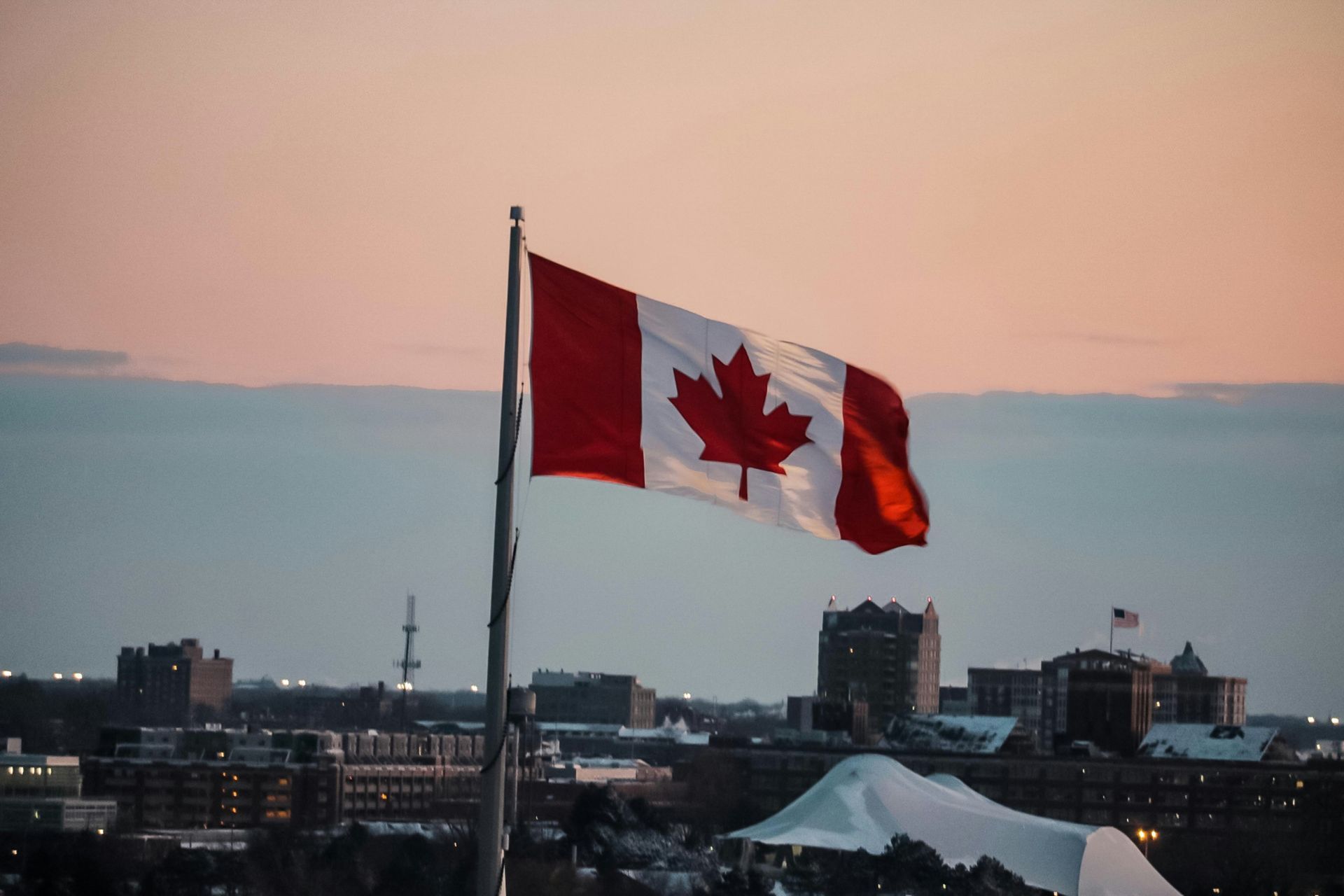 Canadian flag waving in the wind, with a city skyline and sunset sky in the background.