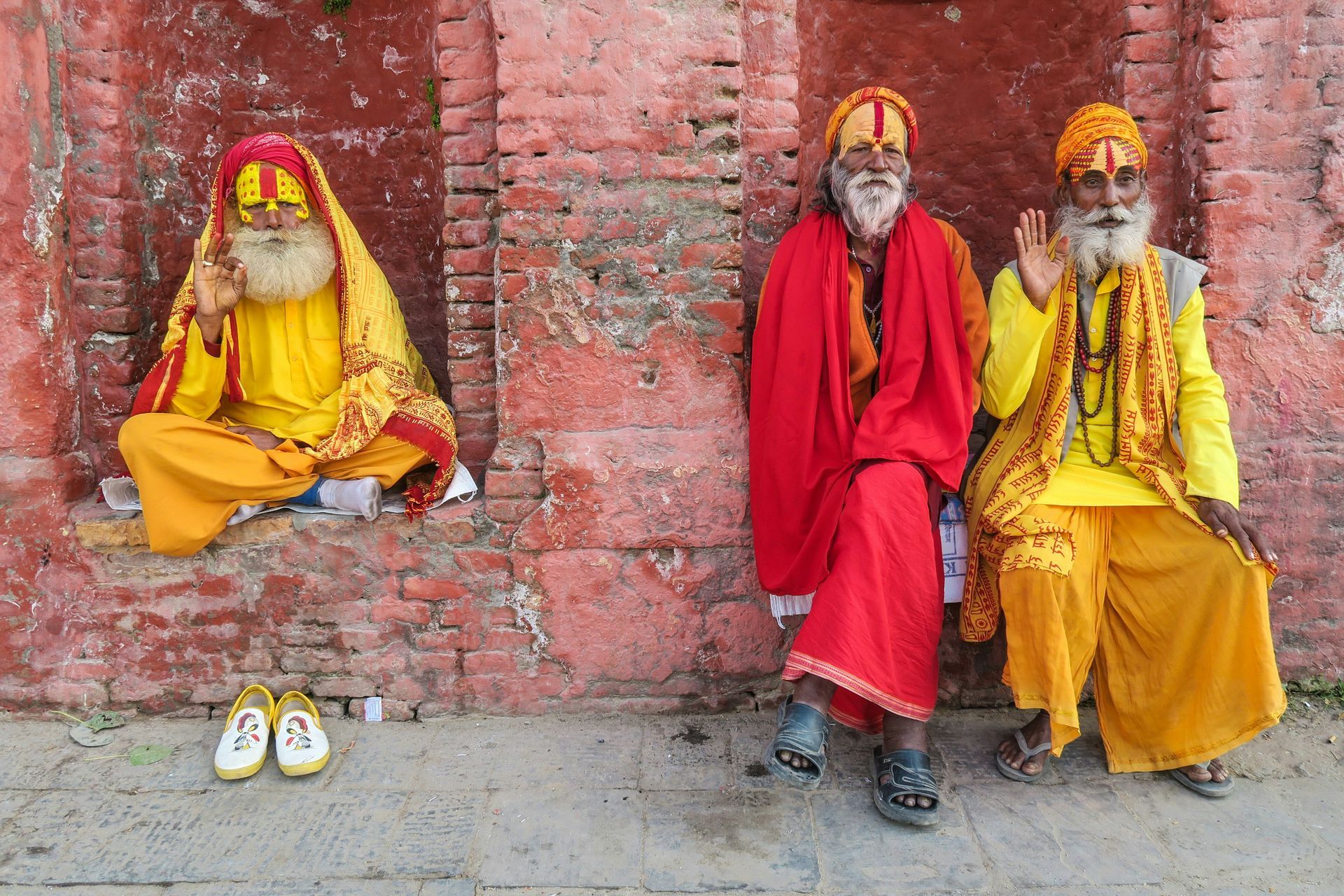 Three robed Nepalese men with long beards and orange headwear seated against a red brick wall, one with hand raised, shoes nearby.