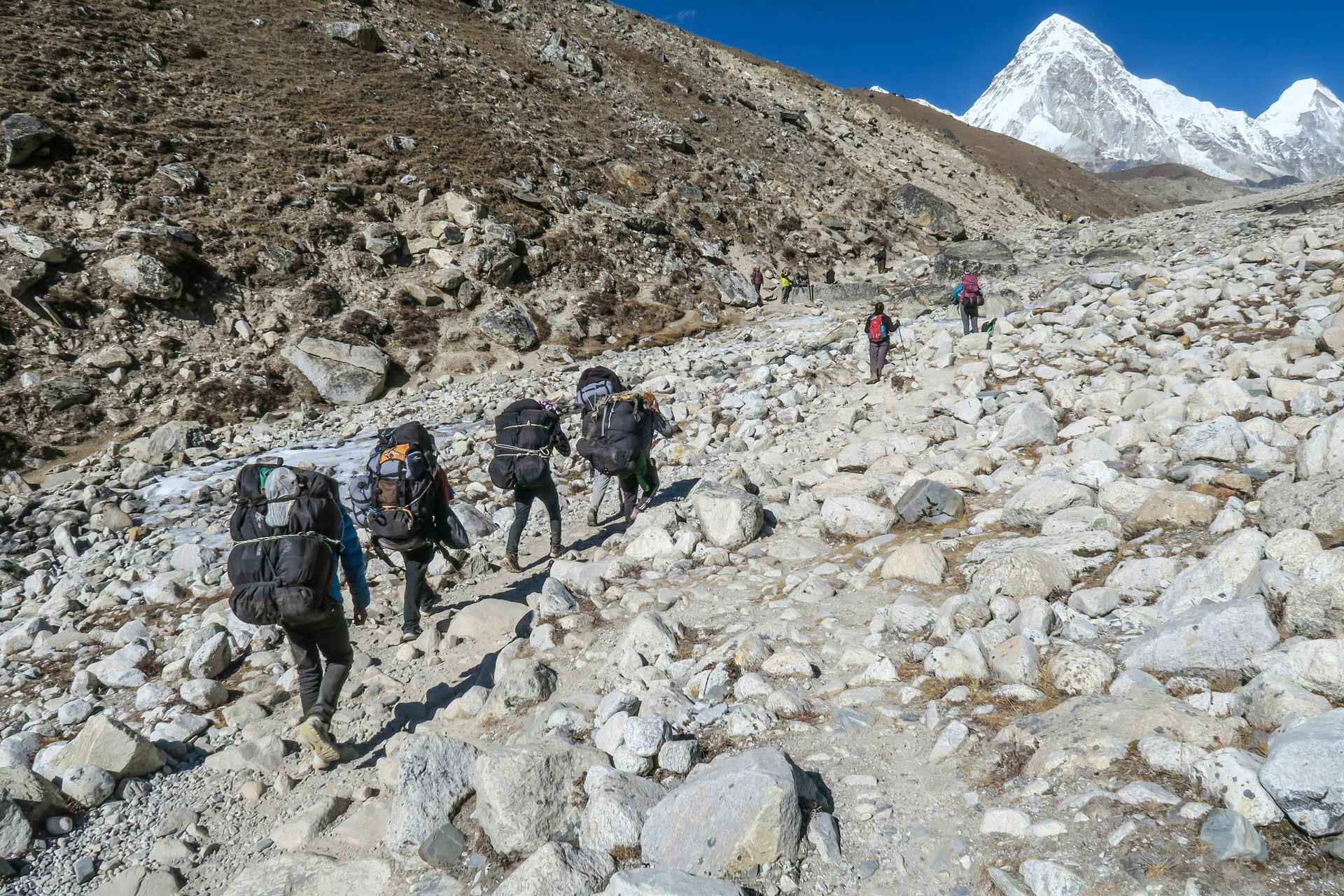 Hikers with large backpacks ascend the rocky Mount Everest trail toward snow-capped mountains in Nepal.