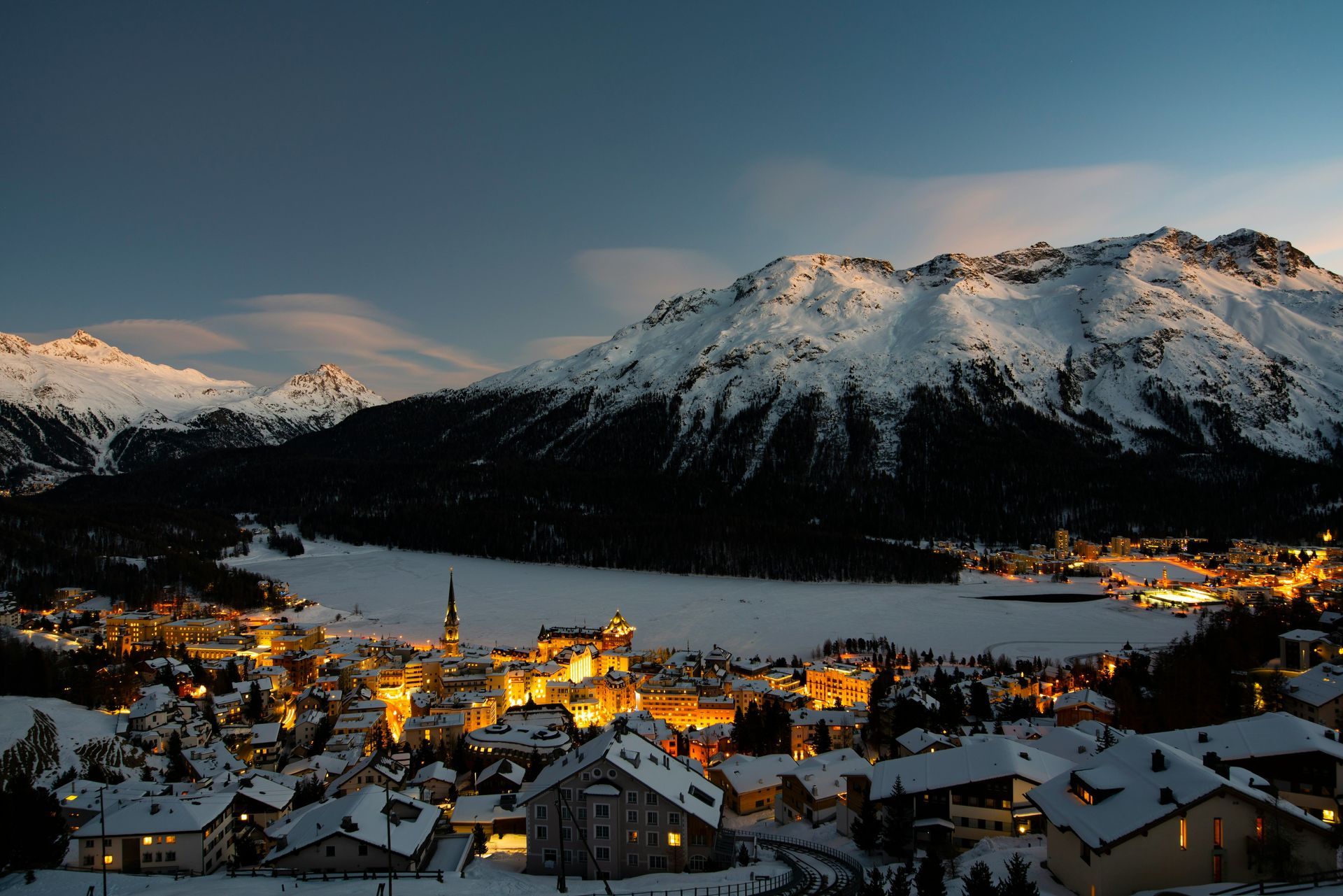 Snowy mountain village of St. Moritz, Switzerland, at dusk, with lights glowing against snow-covered peaks and a dark lake.