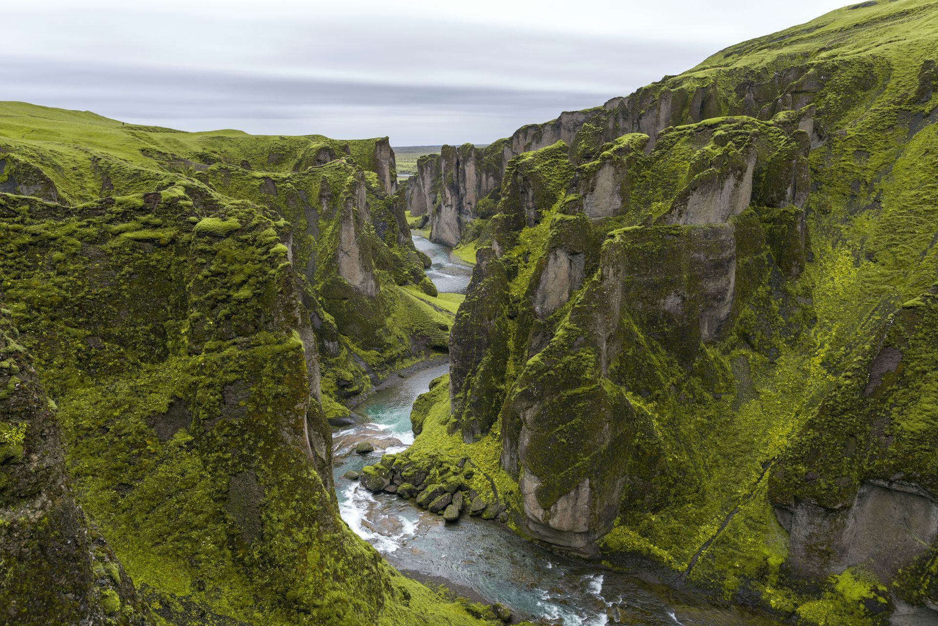 A vibrant, moss-covered canyon with a flowing river under a cloudy sky at Fjaðrárgljúfur, Iceland.