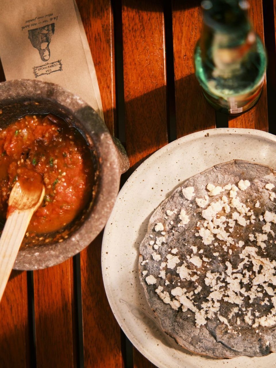 A table setting with a bowl of salsa, a tortilla topped with white cheese, and a bottle in Mexico.
