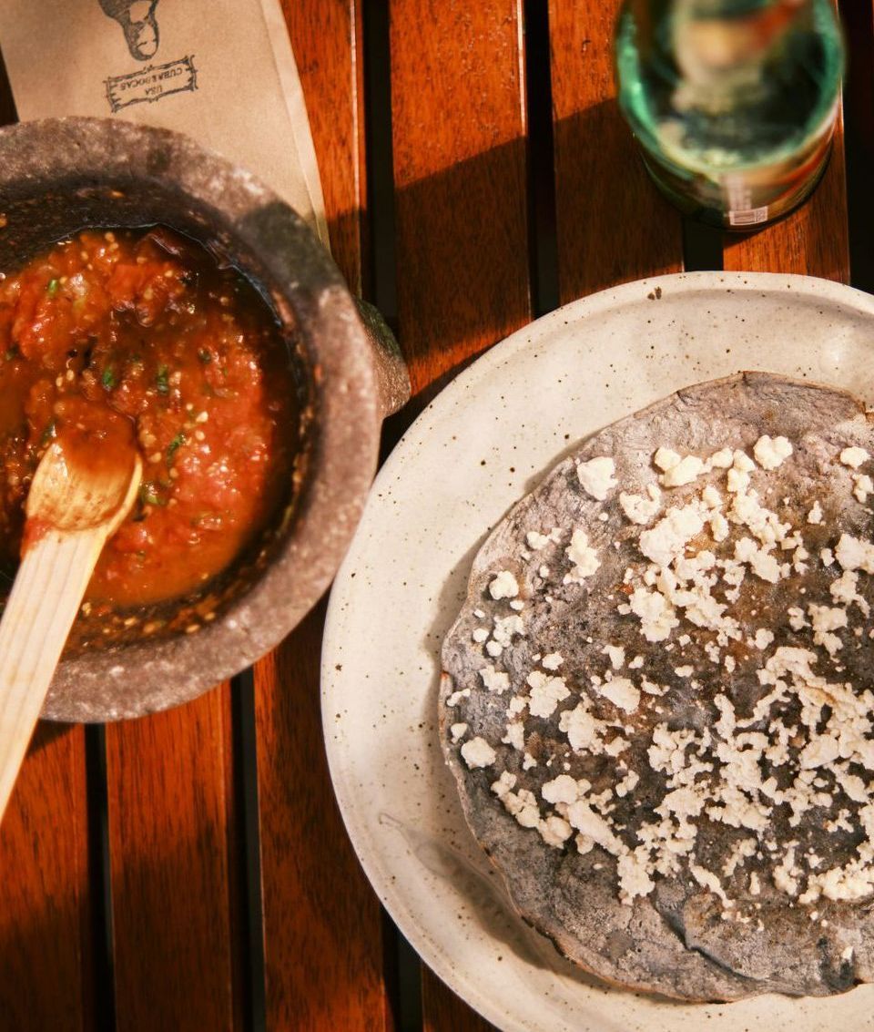 A table setting with a bowl of salsa, a tortilla topped with white cheese, and a bottle in Mexico.