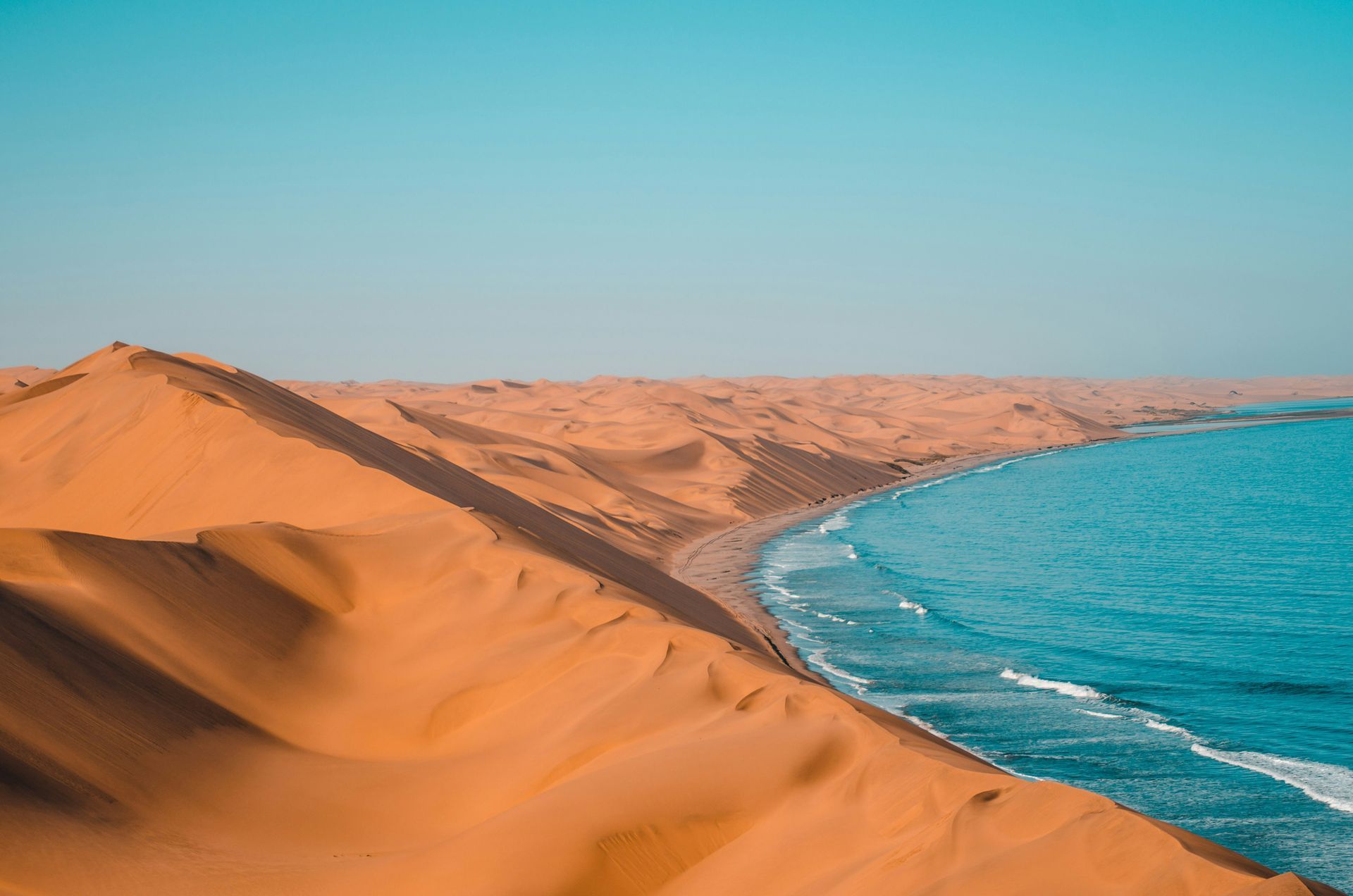 Sandy dunes meeting turquoise ocean under a clear blue sky in Namibia.
