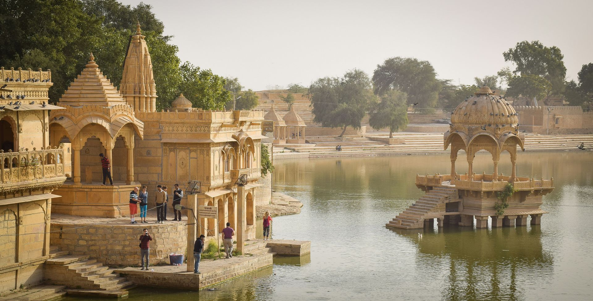 Stone structures line a lake in Jaisalmer, India. People are standing around.