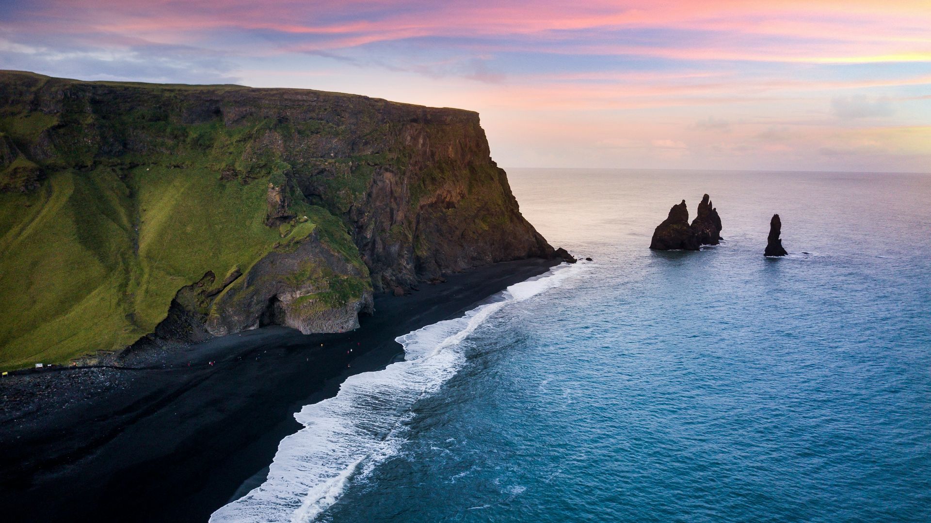The Black sand beach, Reynisfjara Beach, with waves, cliffs, and sea stacks under a colorful sunset sky in Iceland.
