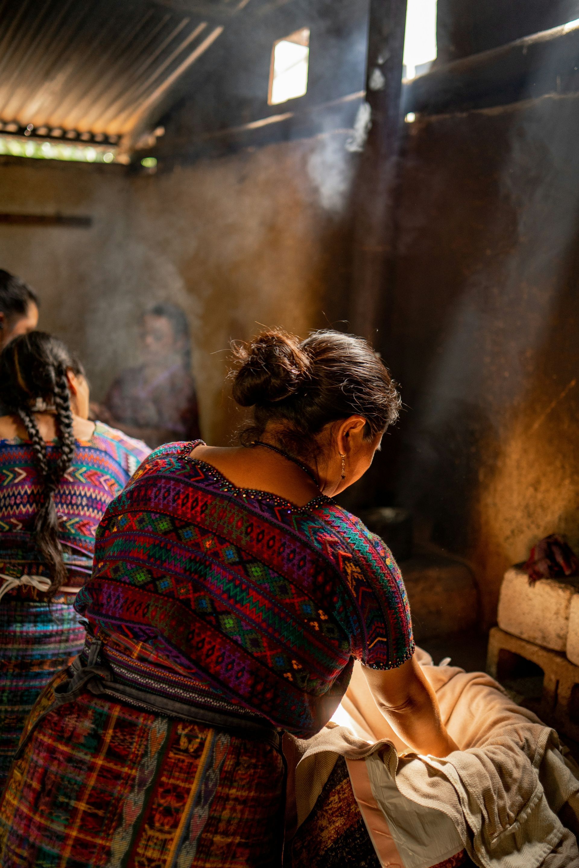 Woman in colorful traditional clothing, stirs a pot in a rustic kitchen, smoke rising, other women nearby in Chile.