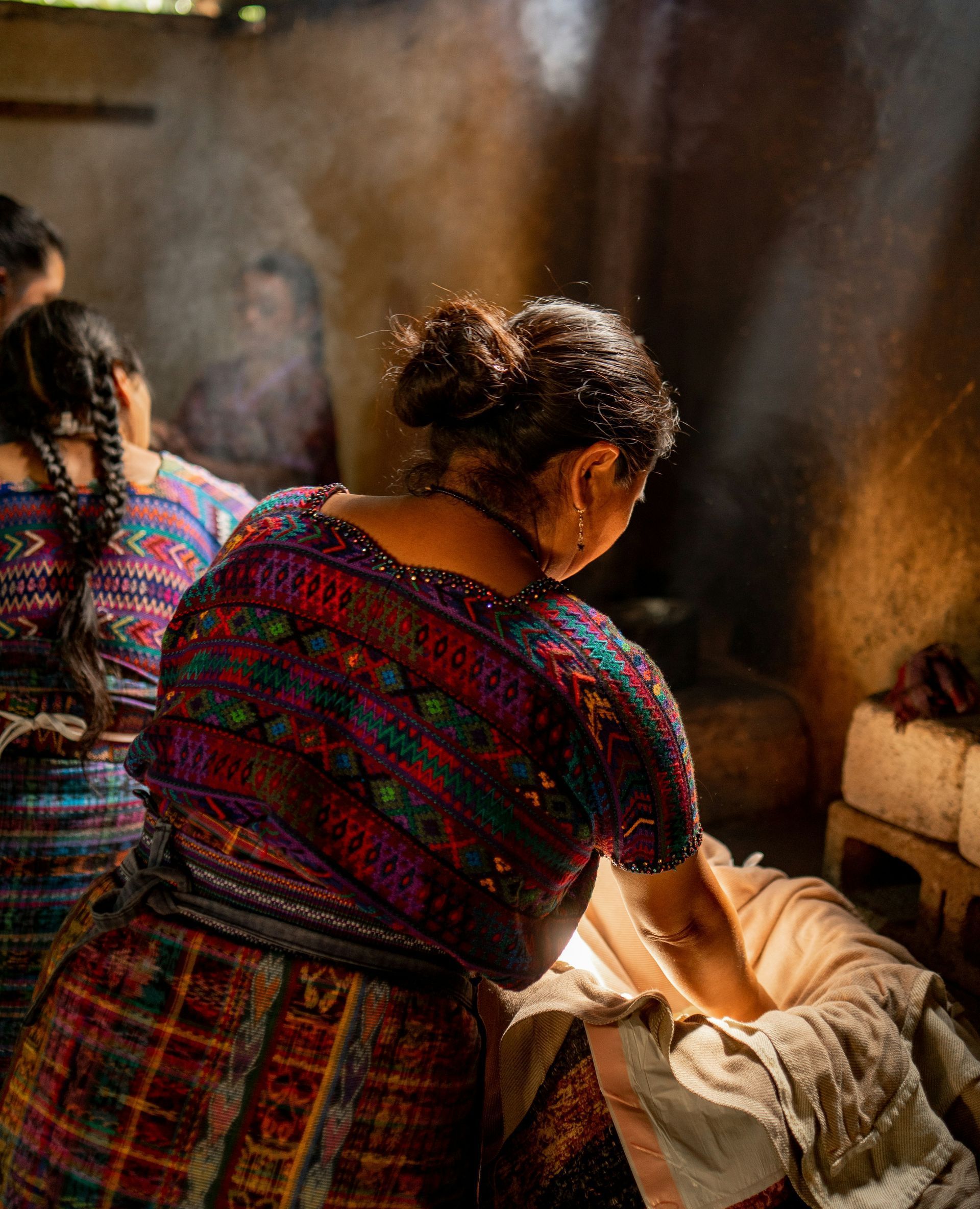 Woman in colorful traditional clothing, stirs a pot in a rustic kitchen, smoke rising, other women nearby in Chile.