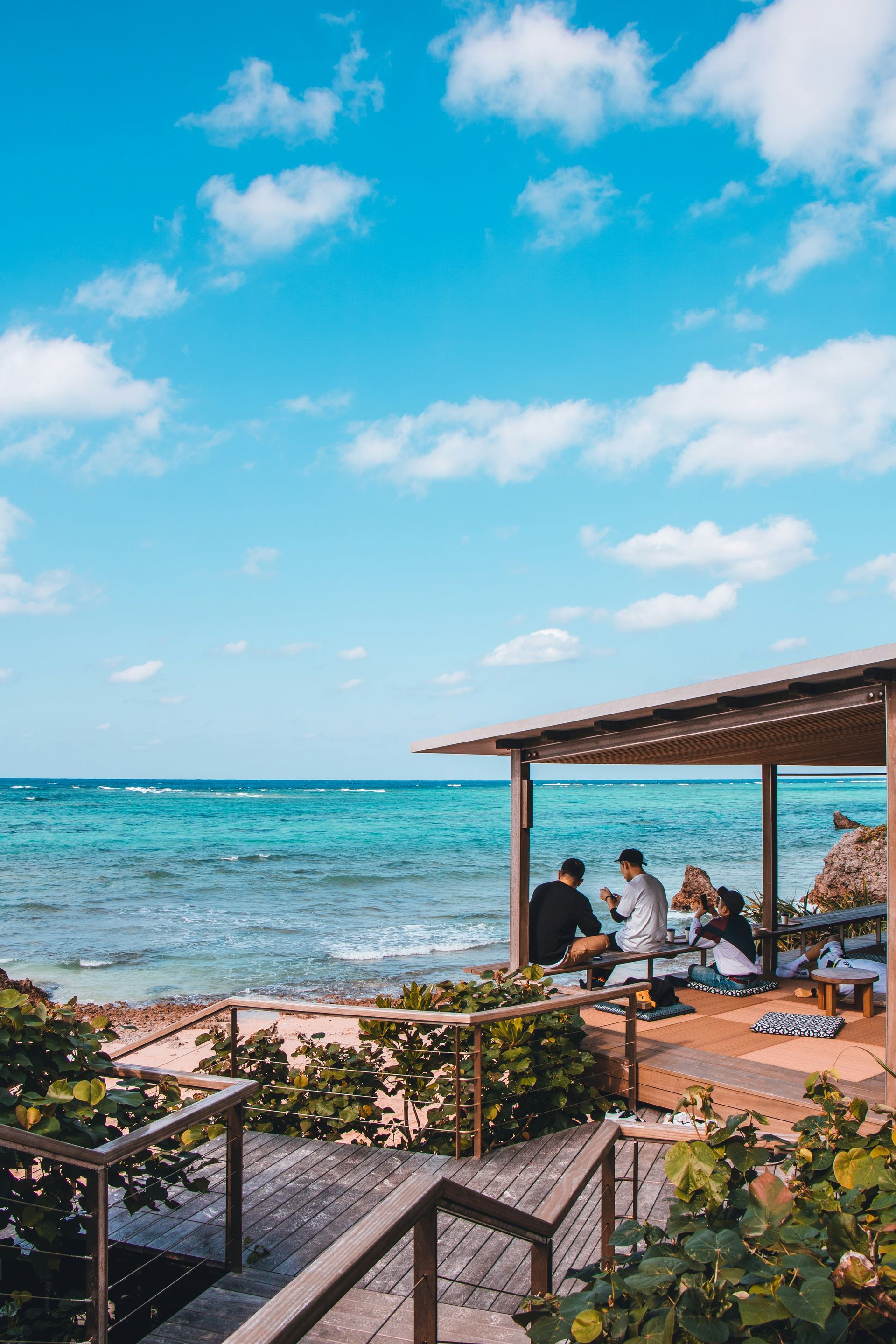 Coastal cafe with turquoise water, blue sky, and people seated under a shaded structure.