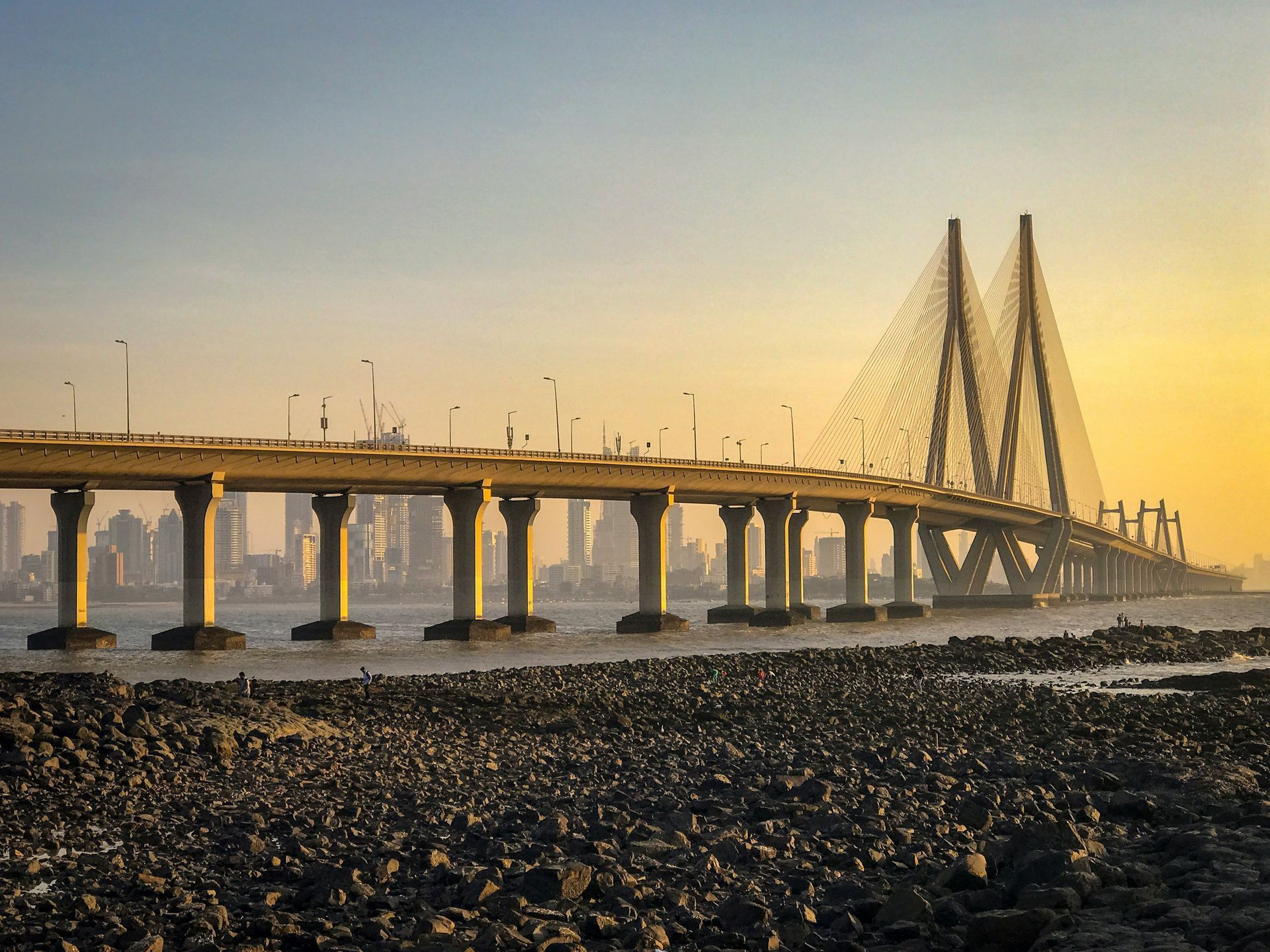 Bridge over water with tall towers, vehicles, and a Mumbai skyline at sunset in India.