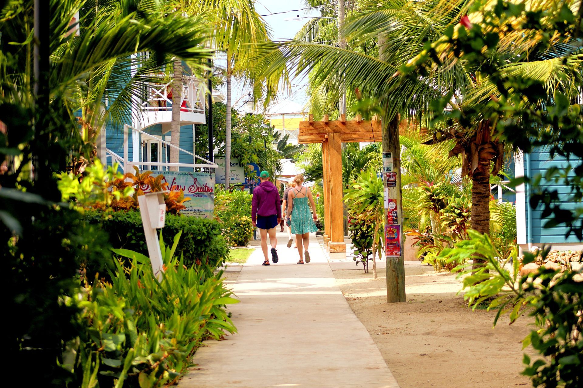Two women walking down a tropical path lined with lush greenery and colorful buildings in Placencia, Belize.