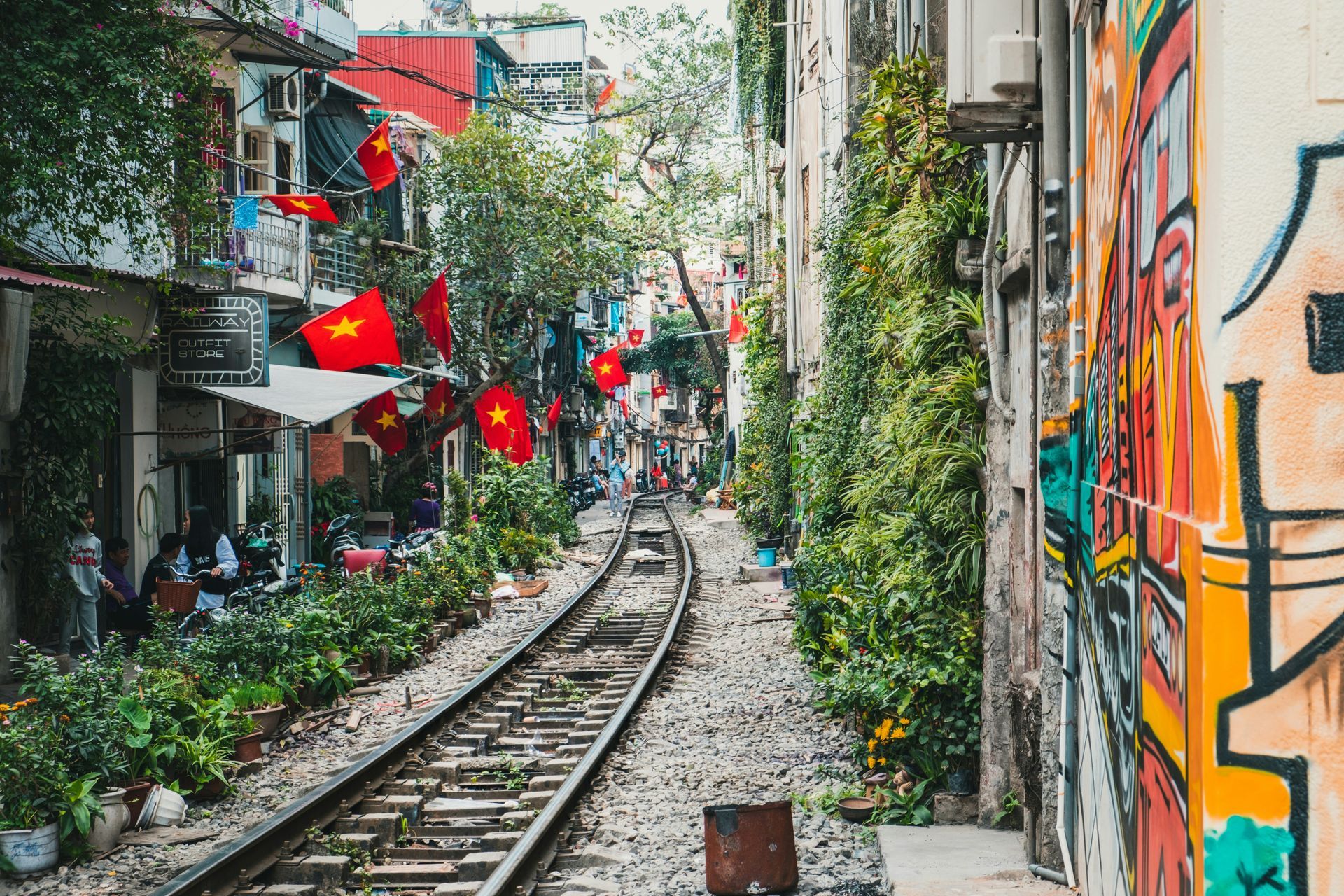 Railroad tracks through a narrow street in Hanoi, Vietnam, flanked by shops, green plants, red flags, and graffiti.