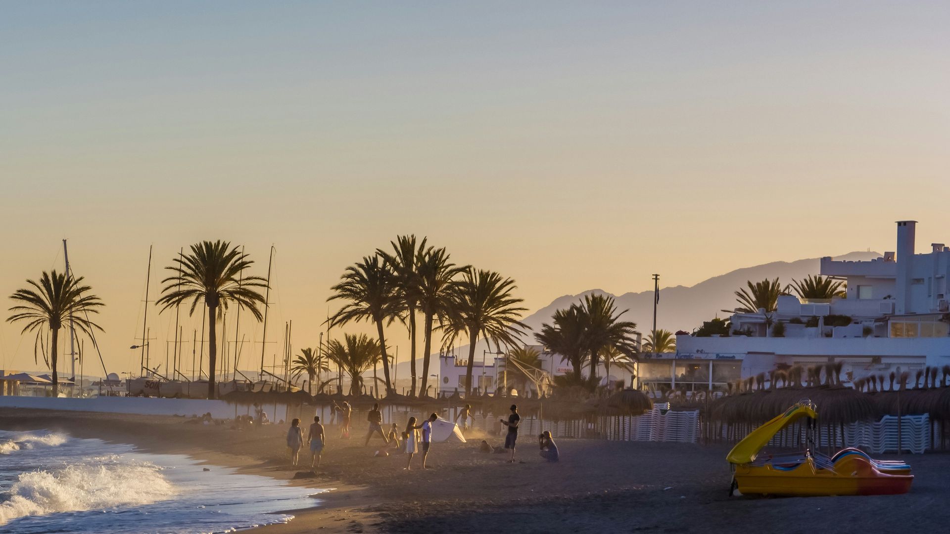 Beach scene at sunset with palm trees, people playing, and buildings in the background in Marbella, Spain.