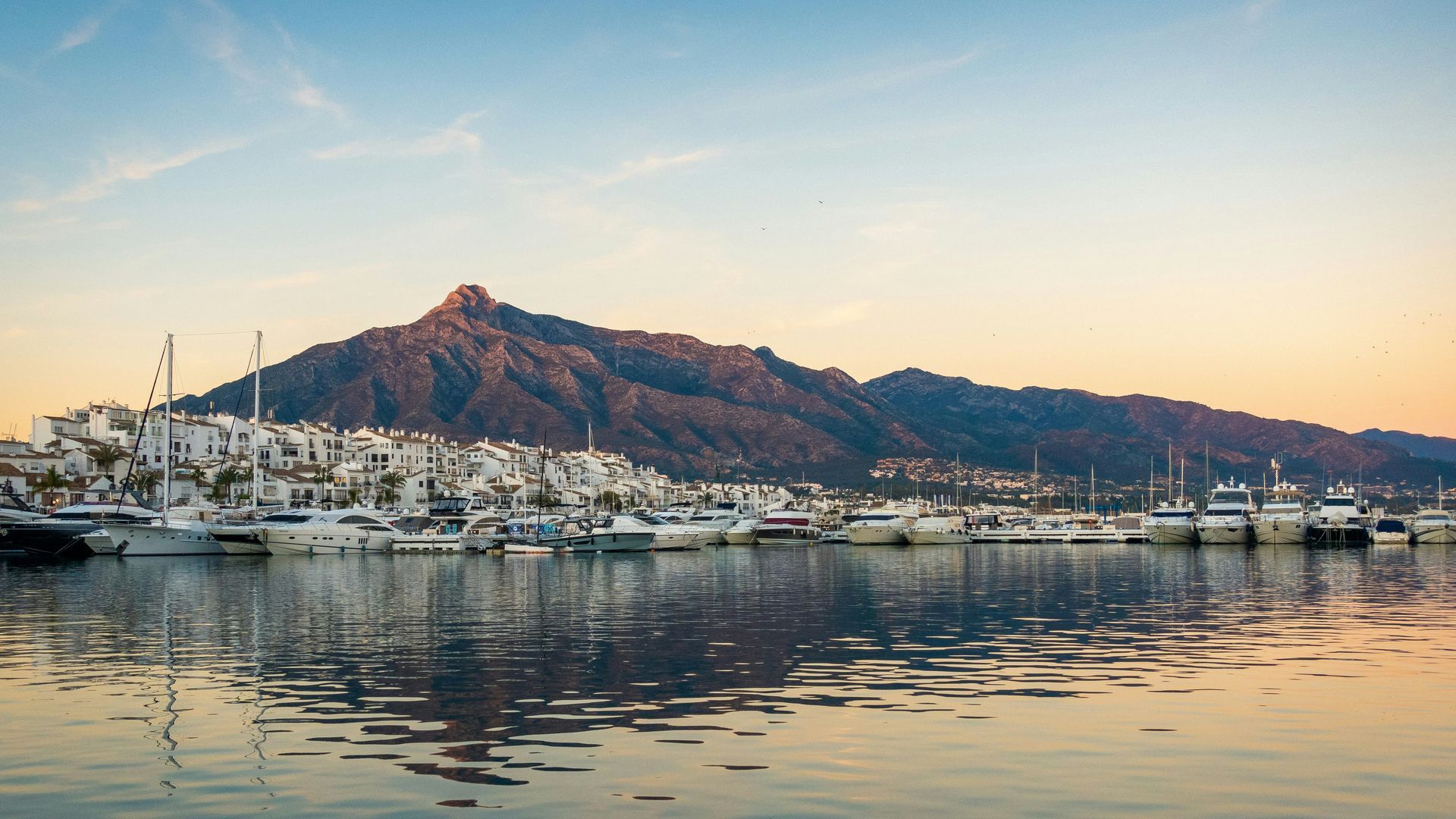 Harbor with boats, white buildings, and a mountain at sunset in Marbella, Spain.
