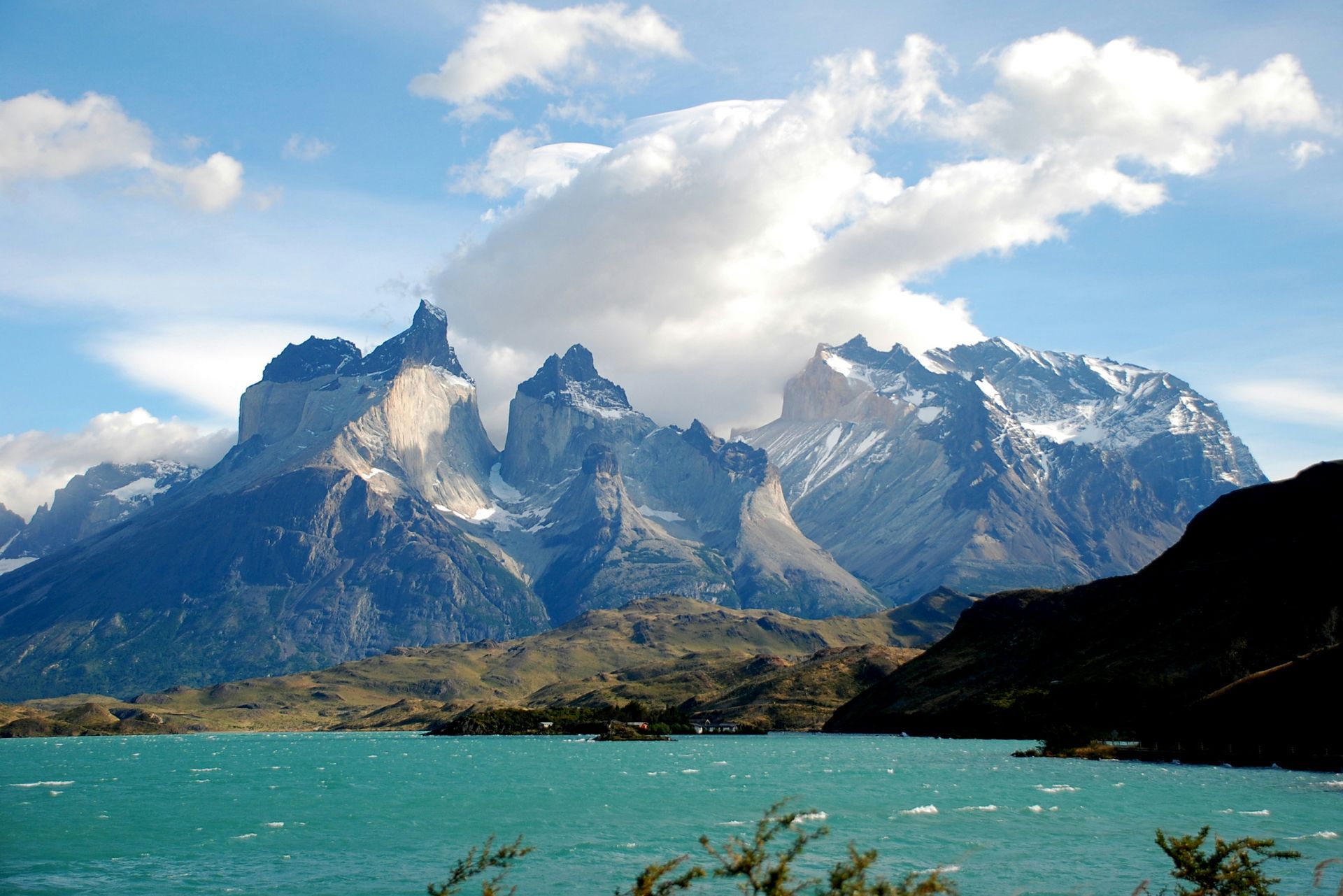 Jagged snow-capped mountains rise above a turquoise lake under a partly cloudy sky in Torres del Paine, Chile.