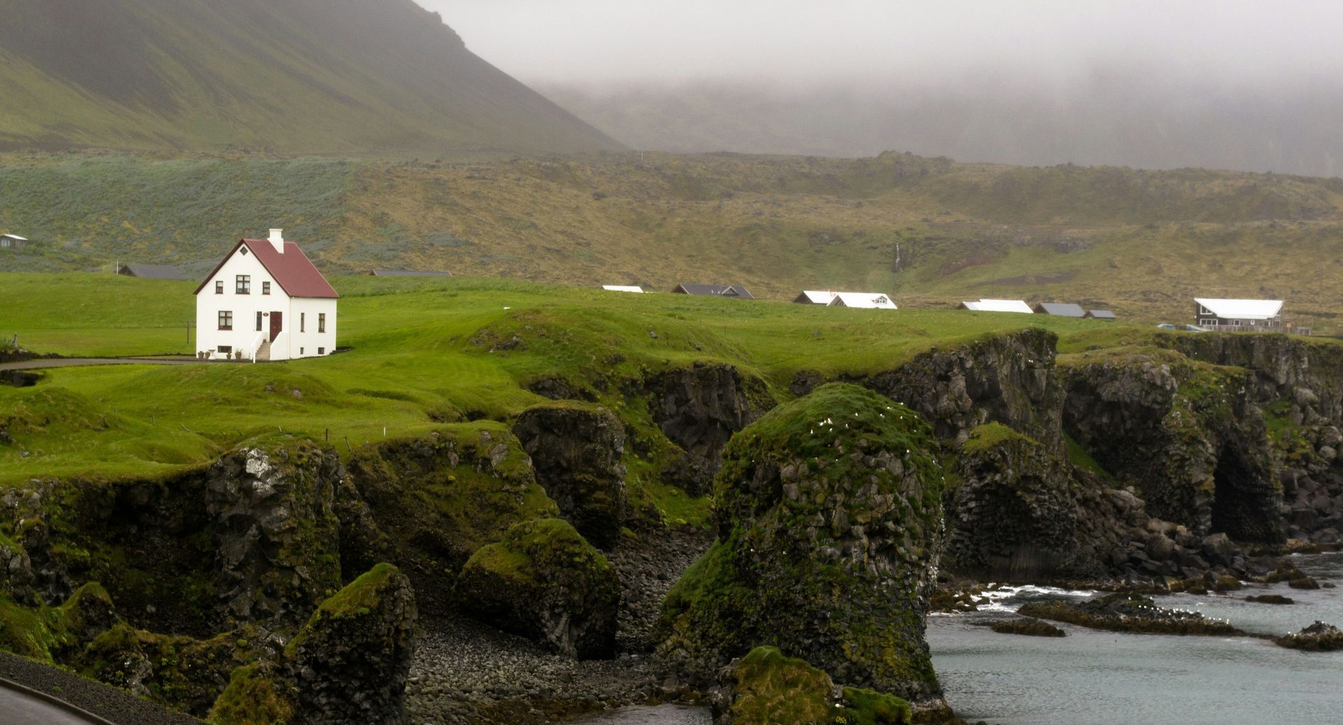 White house on a grassy cliff overlooking a rocky coastline, under a misty mountain in Snæfellsnes, Iceland.