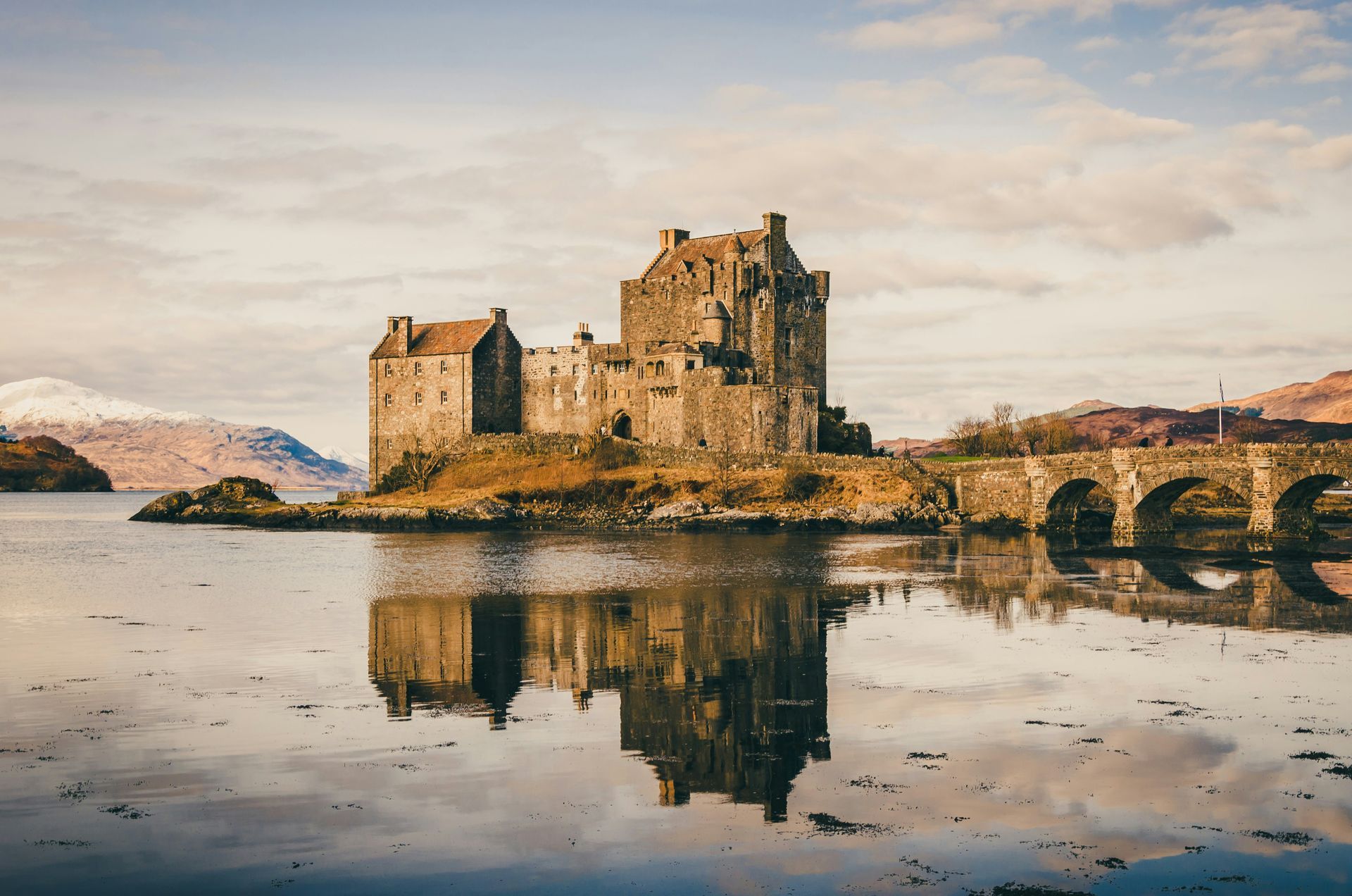 Eilean Donan Castle in Scotland, reflected in calm water. Stone structure with arched bridge, mountains in background.