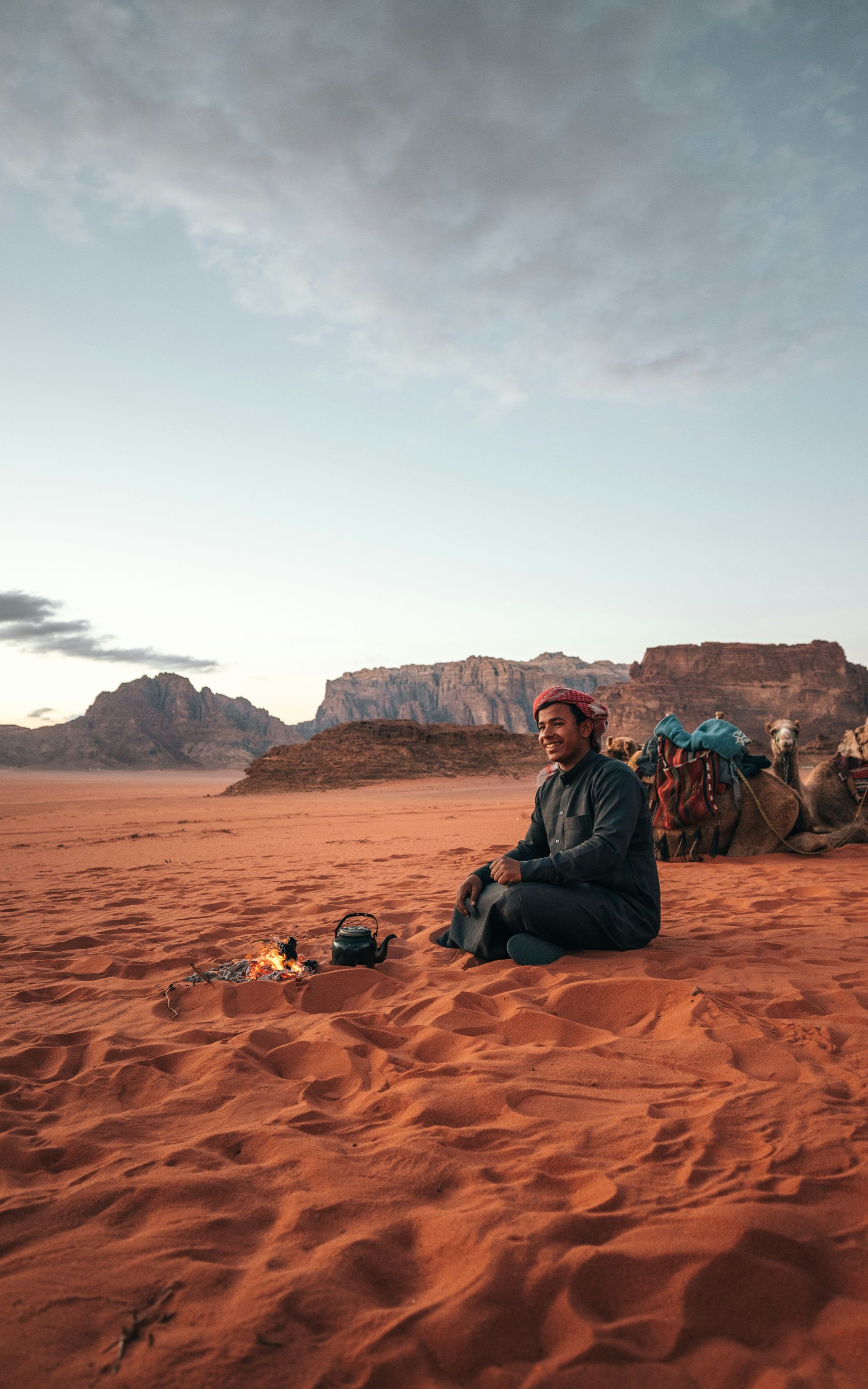Man sits on sand in desert, drinking tea. Mountains in background in Wadi Rum, Jordan.