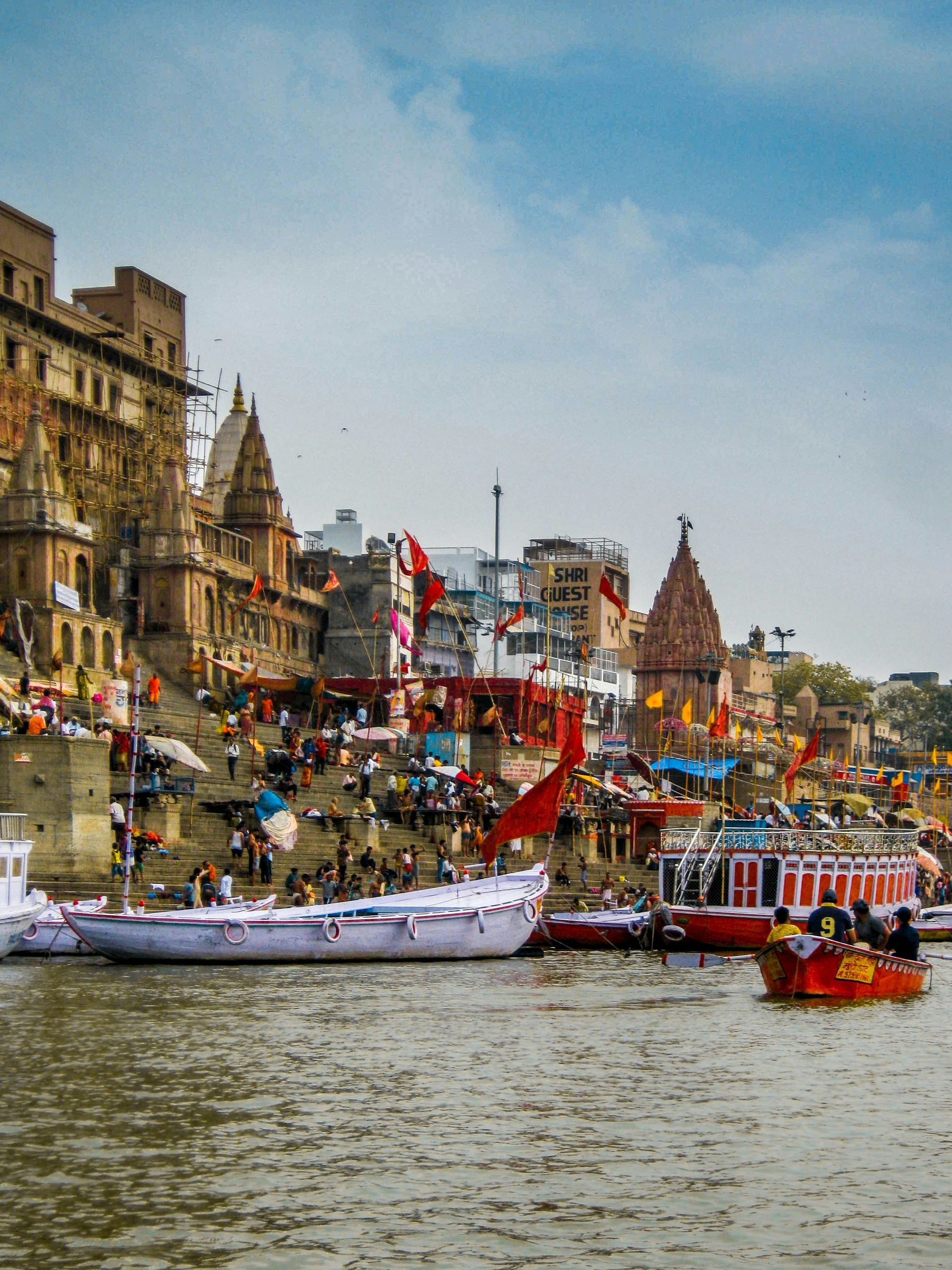 Boats on the Ganges River in Varanasi, India, with people on ghats and colorful buildings under a blue sky.