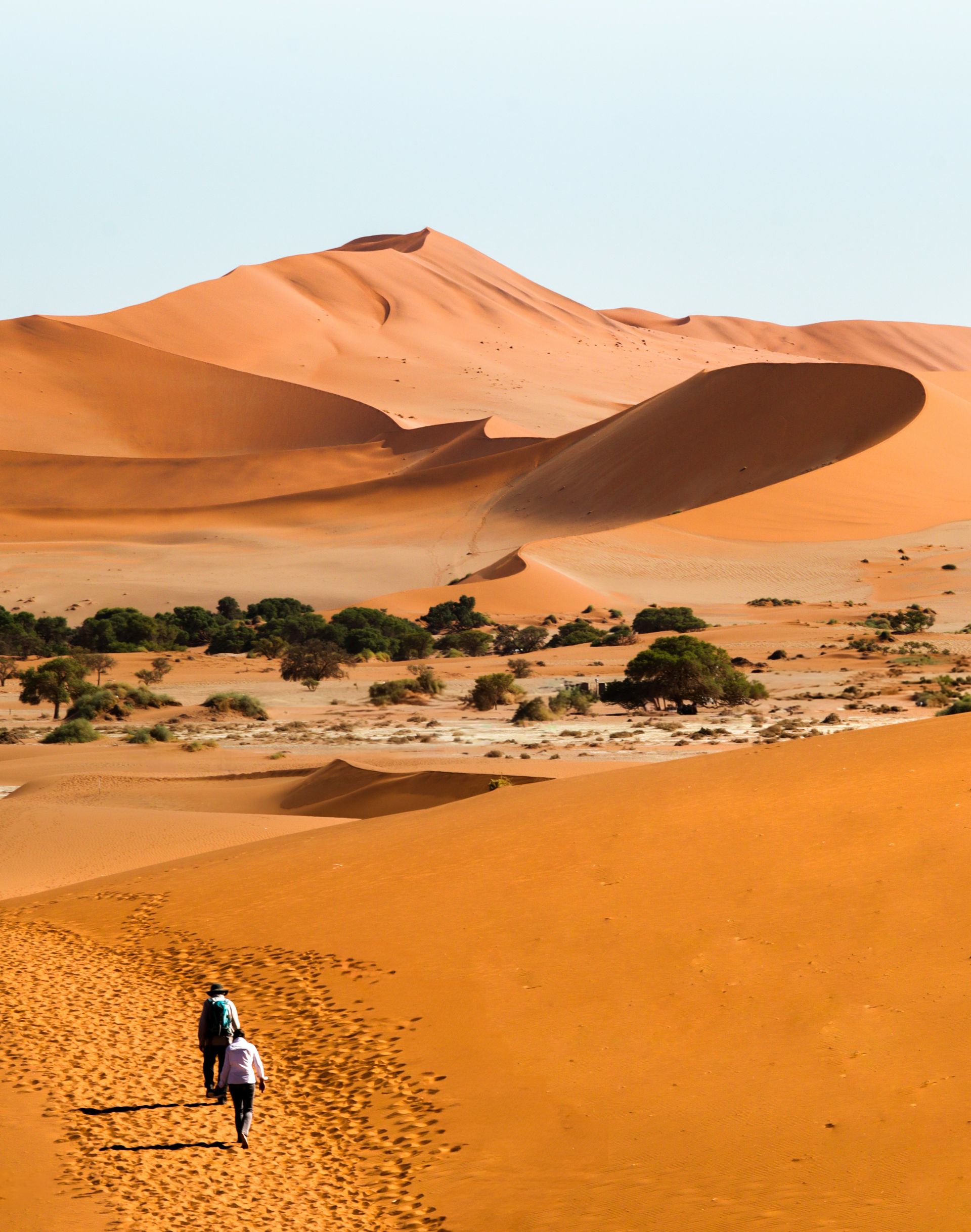 Two people walk up a sand dune towards a larger dune in the Namib Desert, Namibia.