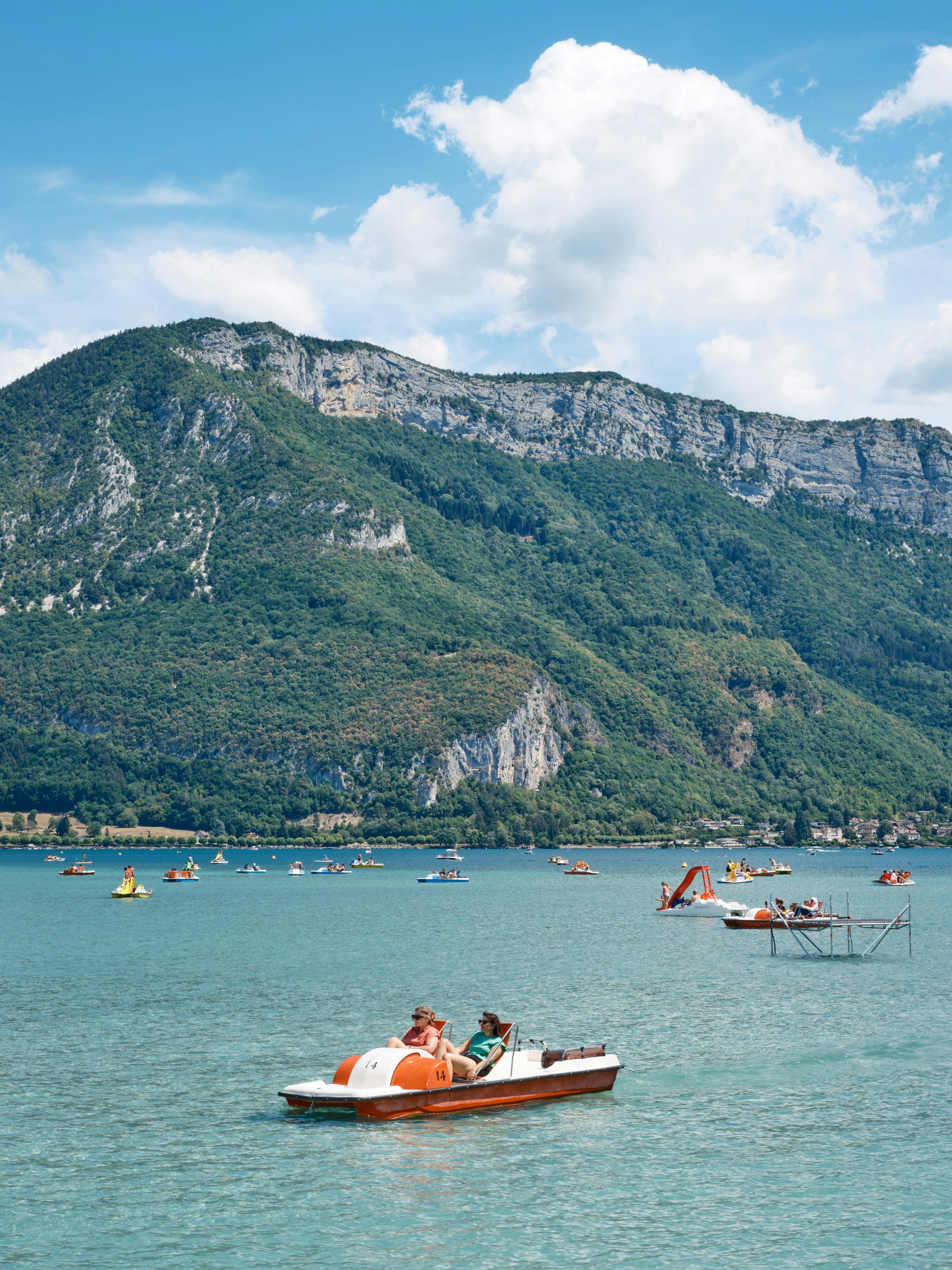A group of people are riding pedal boats on Lake Annecy with mountains in the background in France.