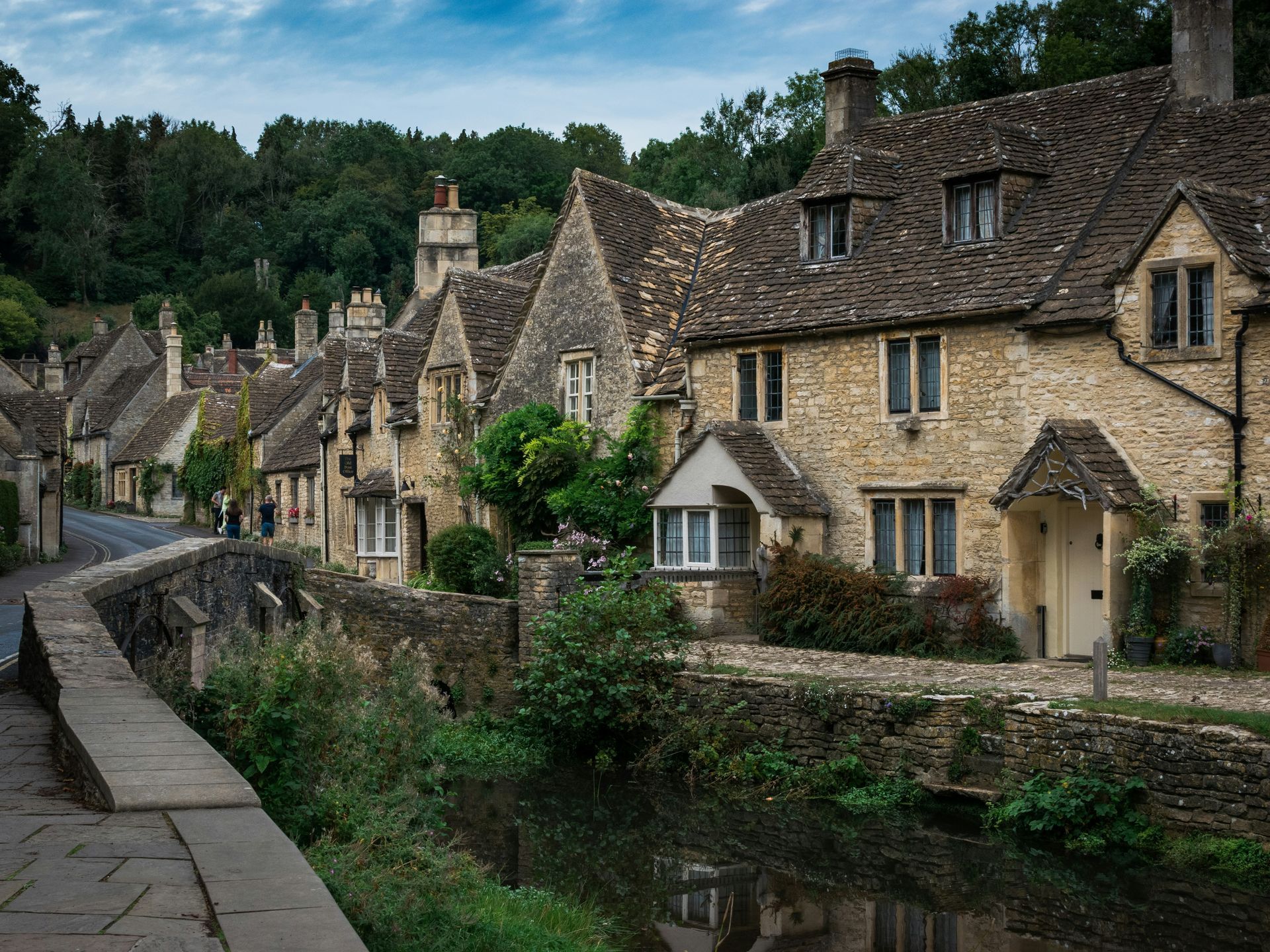 Stone cottages line a street next to a stream in the Cotswolds, England.
