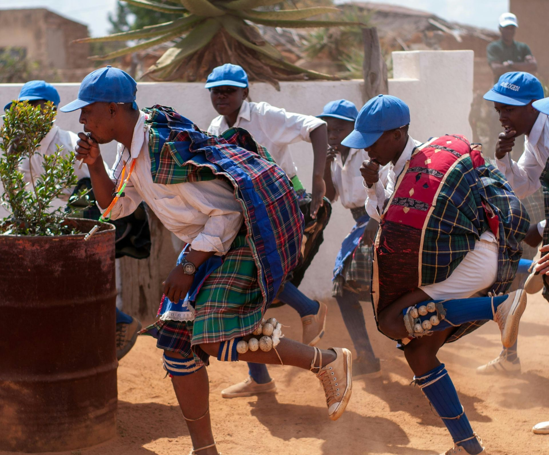 Group of people dancing outdoors in blue caps and traditional clothing in South Africa.