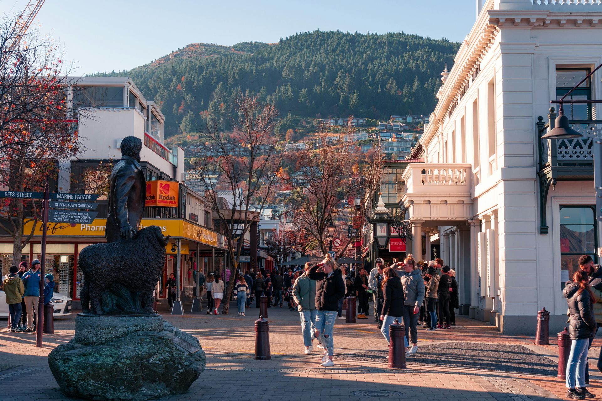A group of people are walking down a street with a statue in the foreground in Queenstown, New Zealand.
