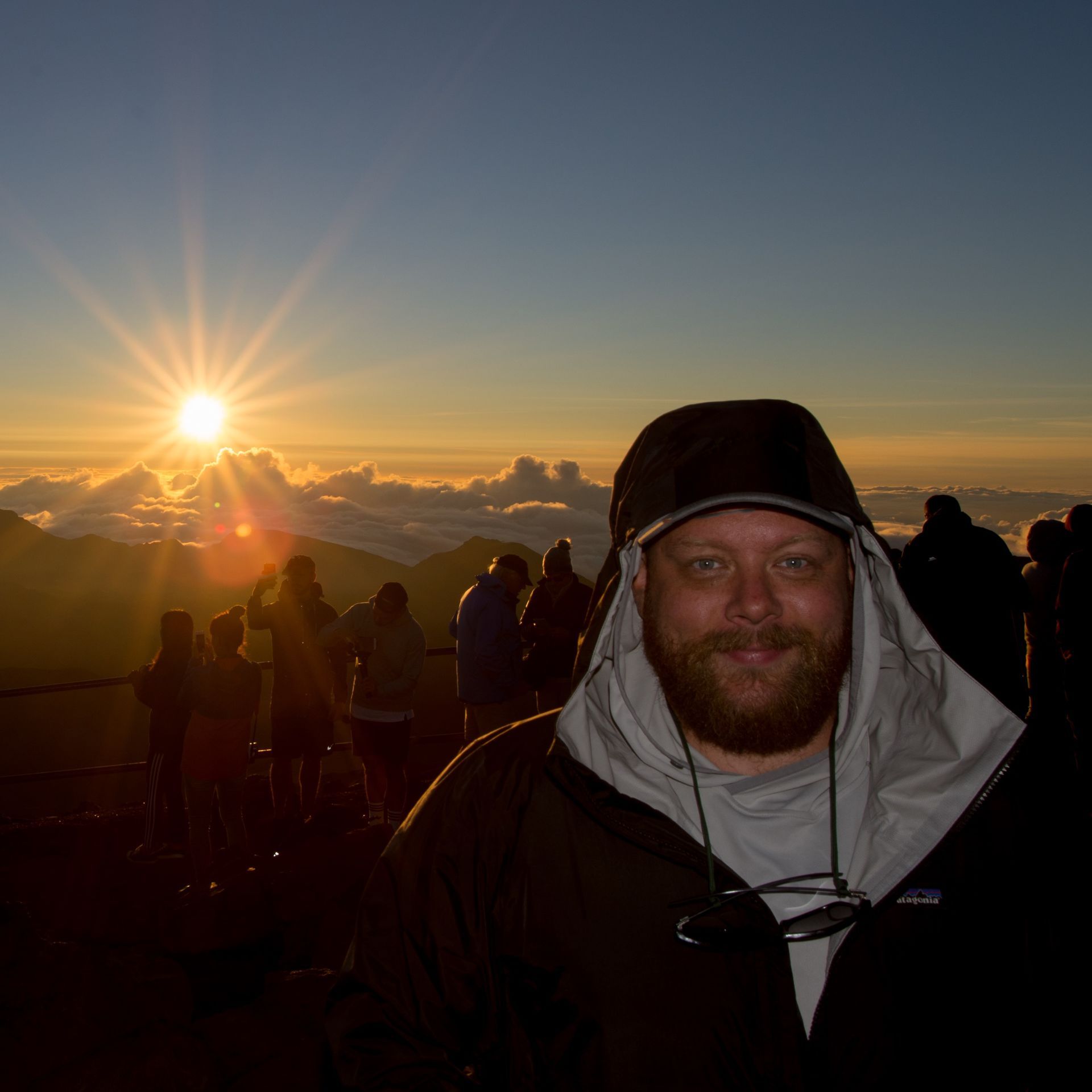Man in a hooded jacket smiles at sunrise over clouds.