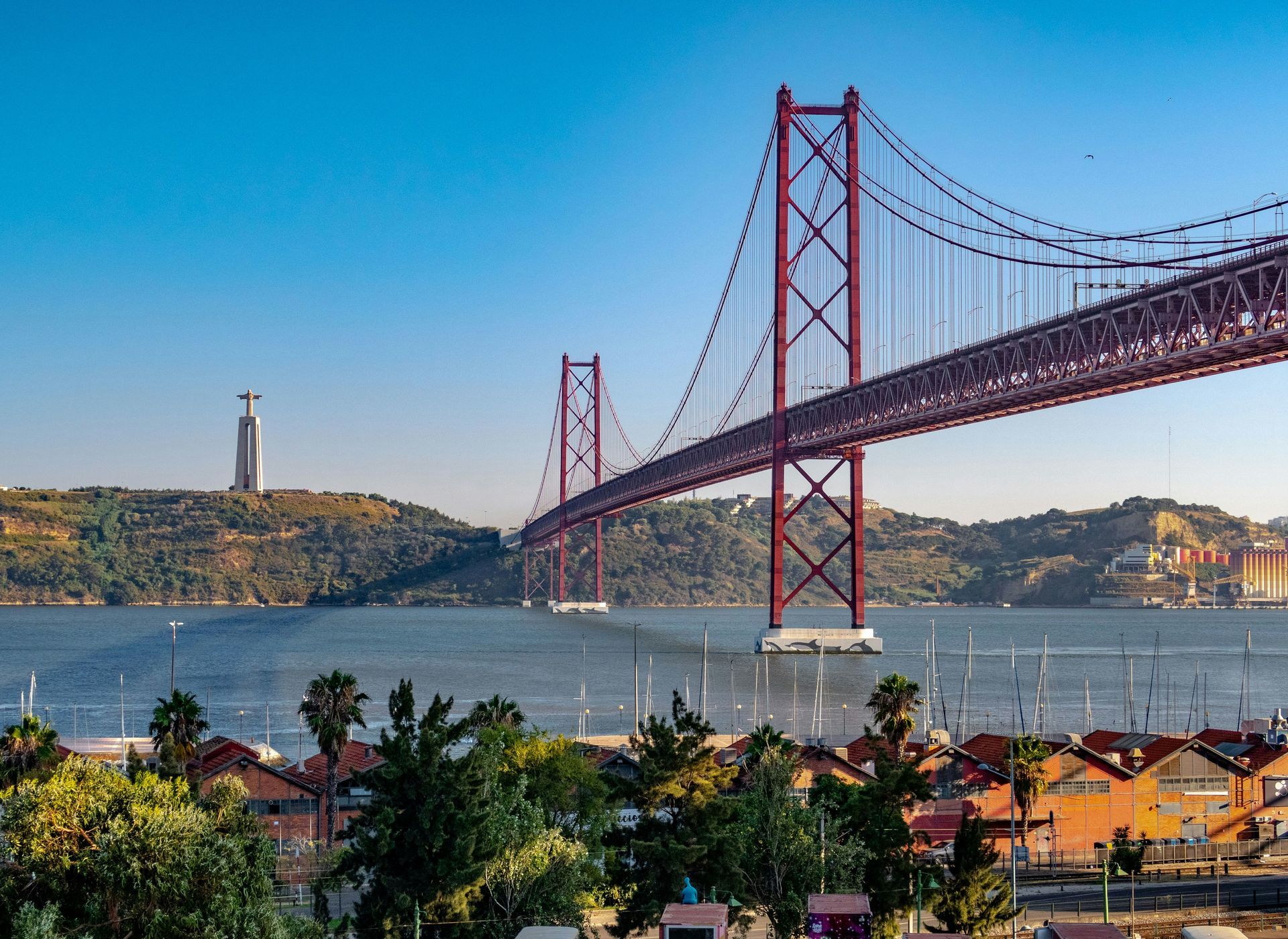 25 de Abril Bridge in Lisbon, Portugal, with Christ the King statue in background; blue sky over water.