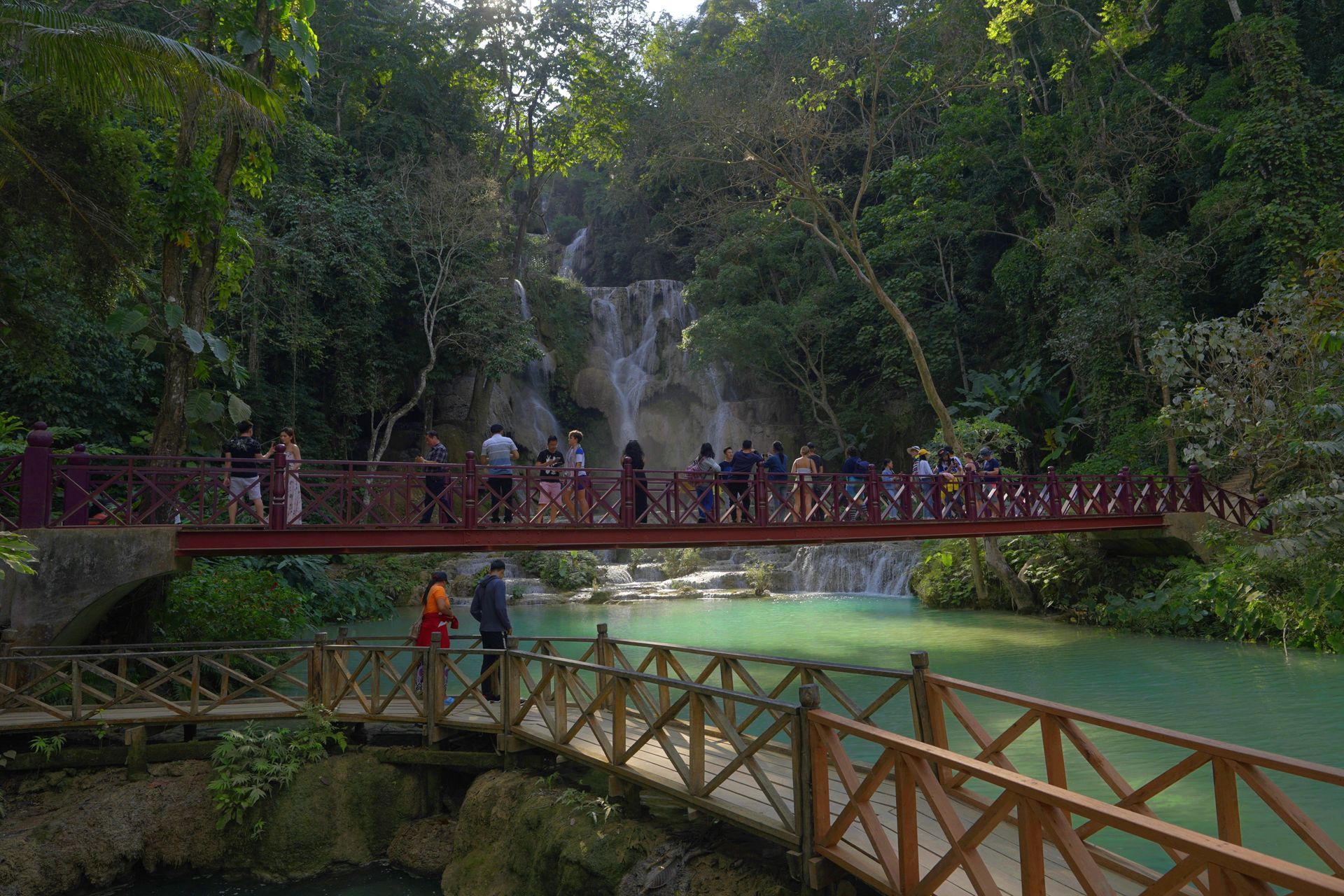 Bridge over turquoise water at Kuang Si Falls in Laos. Tourists gather. Waterfall in the background, surrounded by greenery.