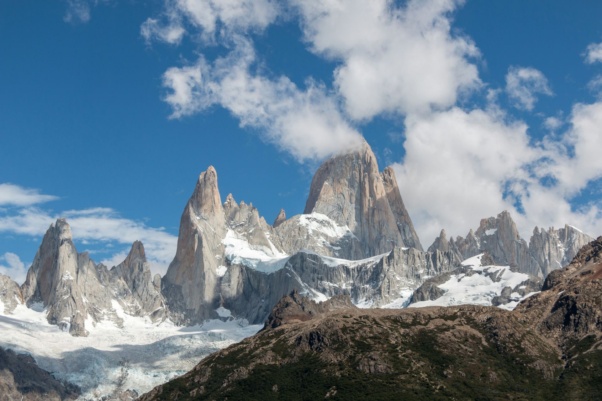 Jagged, snow-capped mountains rise against a blue sky with fluffy white clouds in El Chaltén, Argentina.